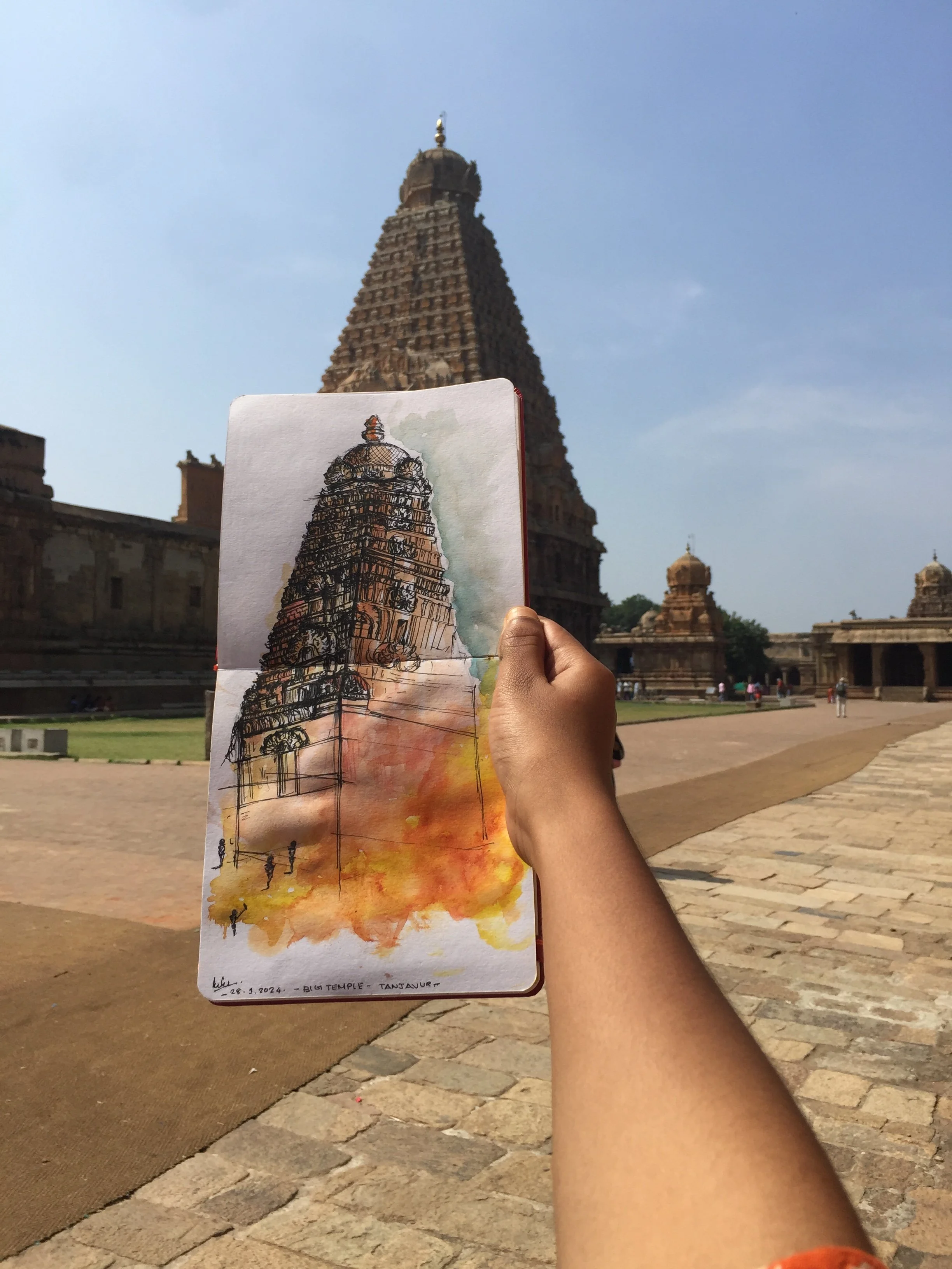 A hand holding a watercolor painting of the Brihadeeswarar Temple, Tanjore, with the actual temple in the background under clear skies.