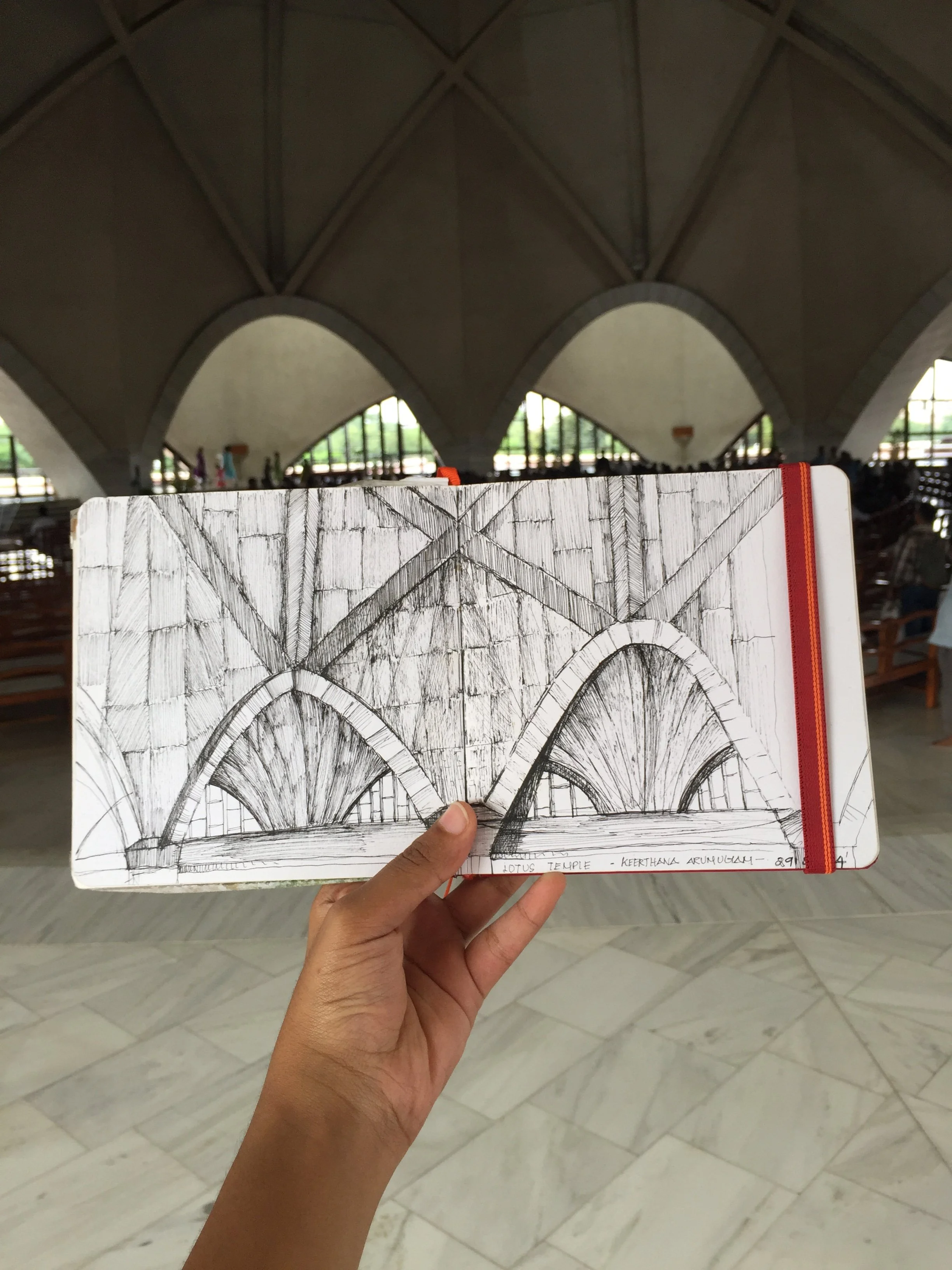 A hand holding a sketchbook featuring a pencil drawing of an architectural structure with multiple arches inside a large building. The background shows a spacious interior with curved ceiling and large windows, likely a temple or similar structure.