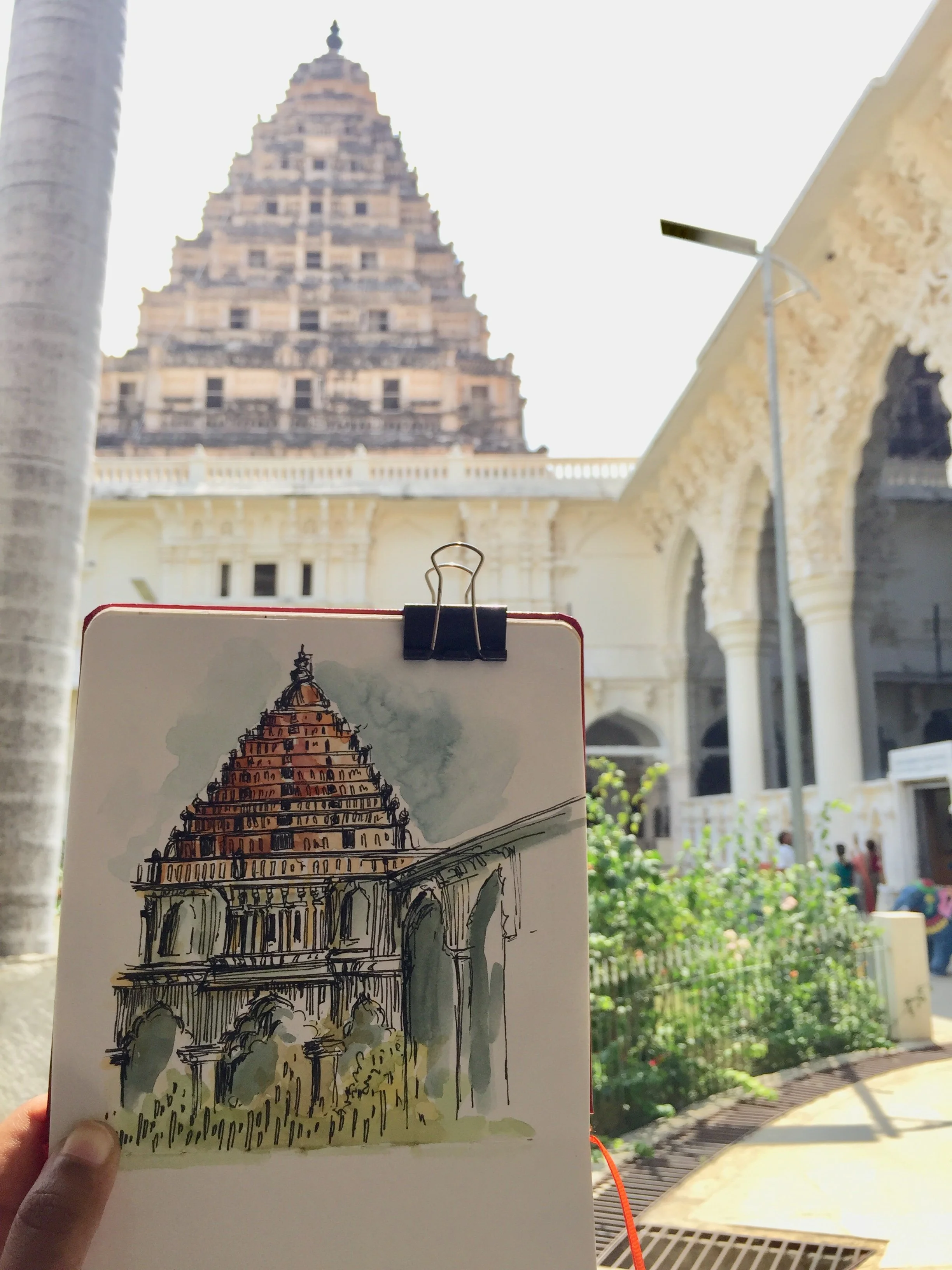 Drawing of a tiered temple structure, depicted in an urban setting with a historic building in the background, taken outdoors with sunlight.
