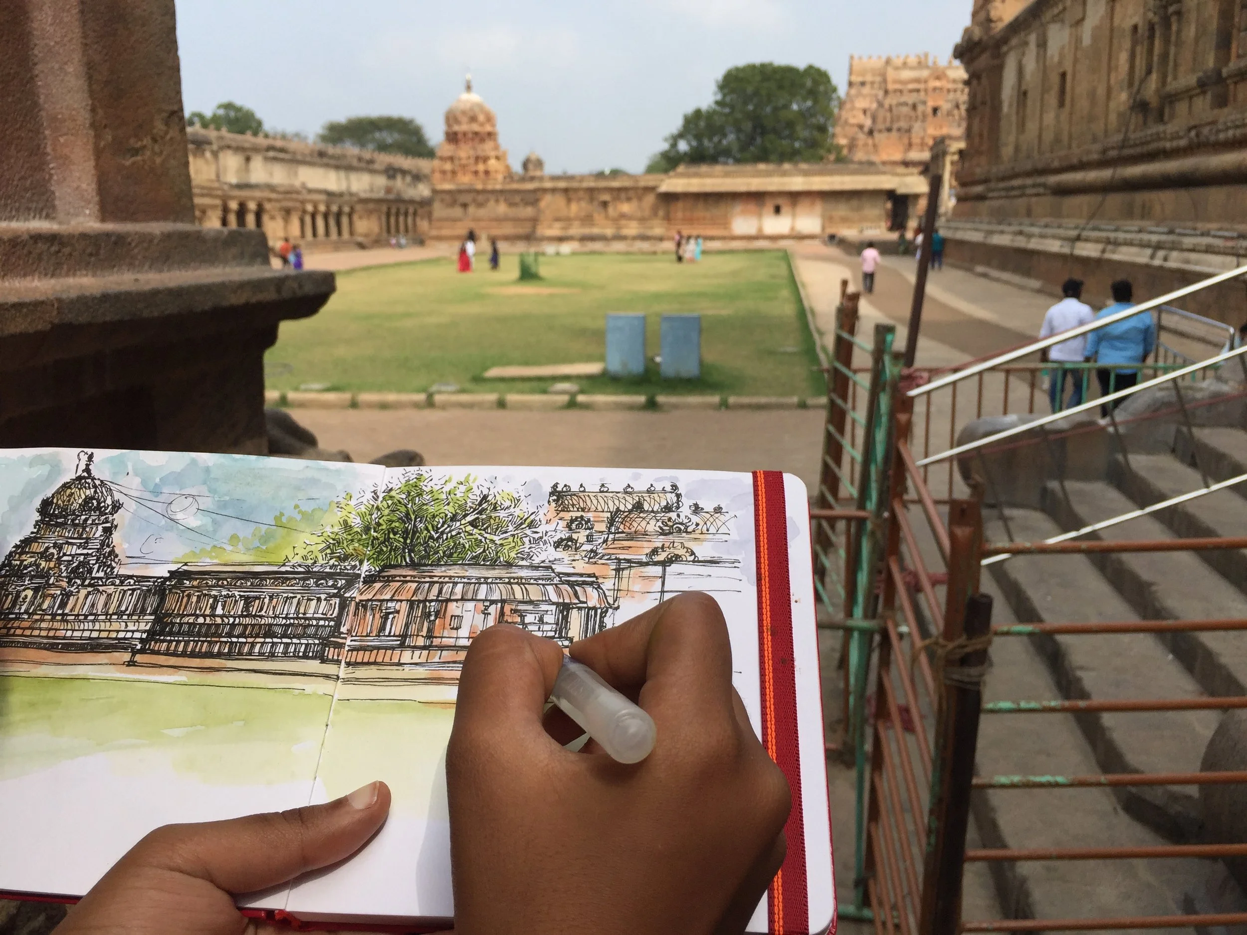 A person holding a sketchbook with a drawing of a historic building, with a background of ancient Indian architecture and a courtyard with visitors.