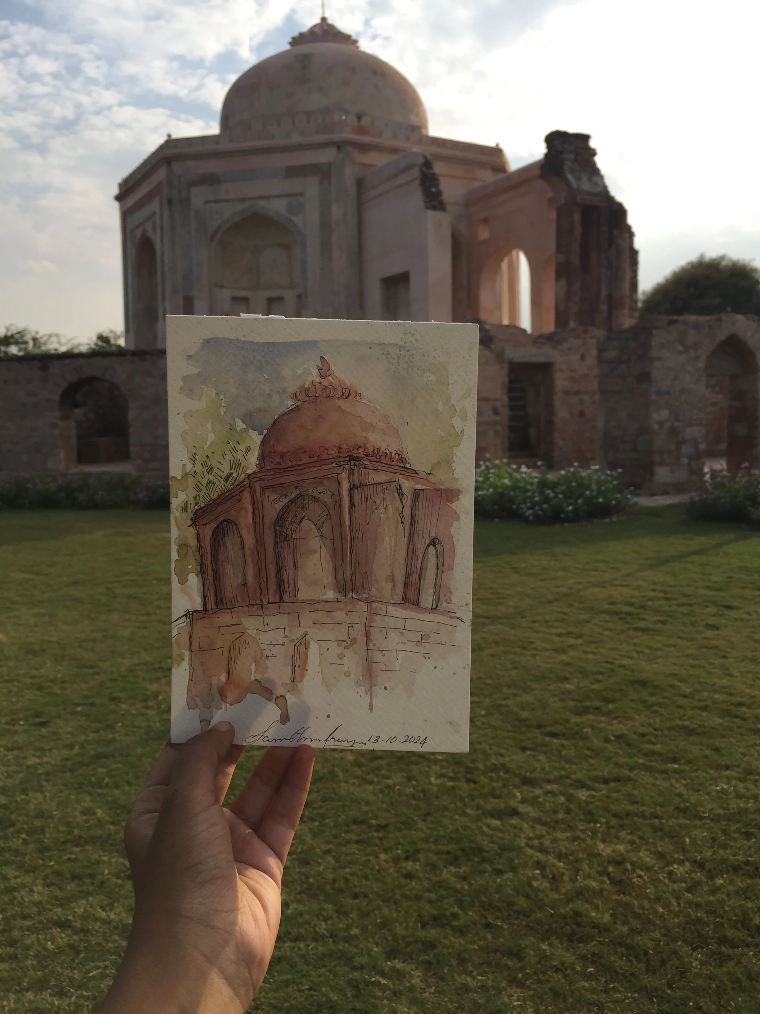 A watercolor painting of a domed monument held in front of the actual ruined monument in the background, with cloudy sky and green grass.