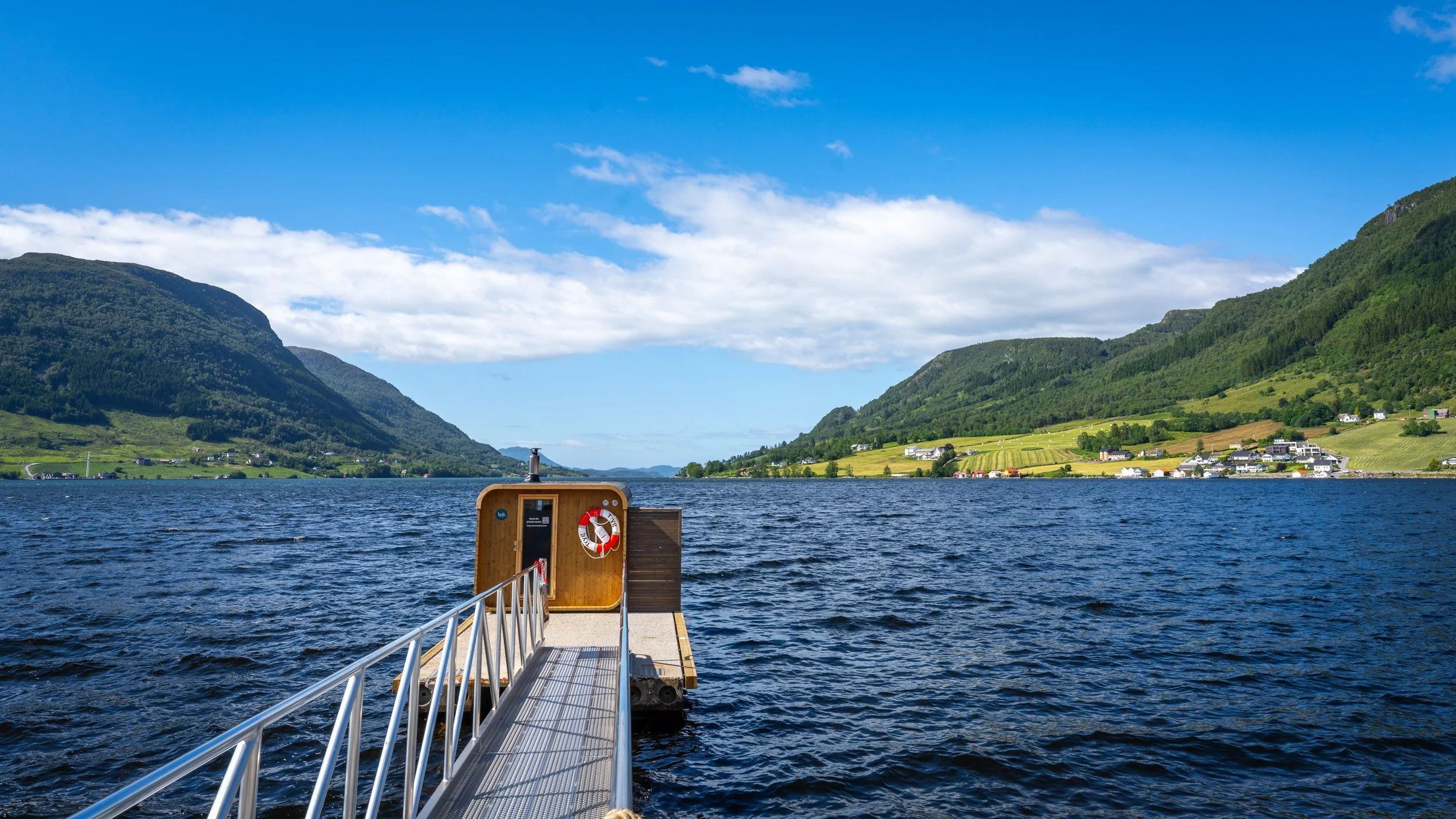 Heit Ølen. Utsikt fra flytebrygge over en fjord omgitt av grønne fjell og landsbyer med hus, under blå himmel med noen skyer.