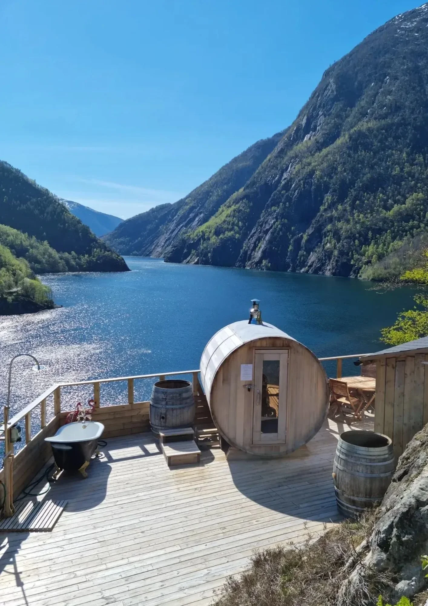 Heit Åkrafjorden. Utsikt over en fjord omgitt av fjell med grønne trær, sett fra en treterrasse med boblebad og en liten sauna laget av tre.