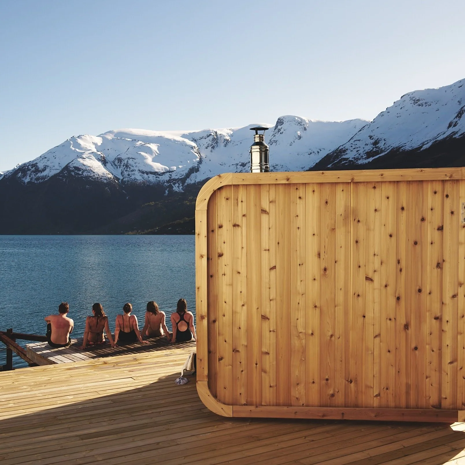 Heit Grimo, Hardangerfjorden, Sørfjorden. Fire personer som sitter på en brygge av tre foran en stor fjord, med snødekte fjell i bakgrunnen. 