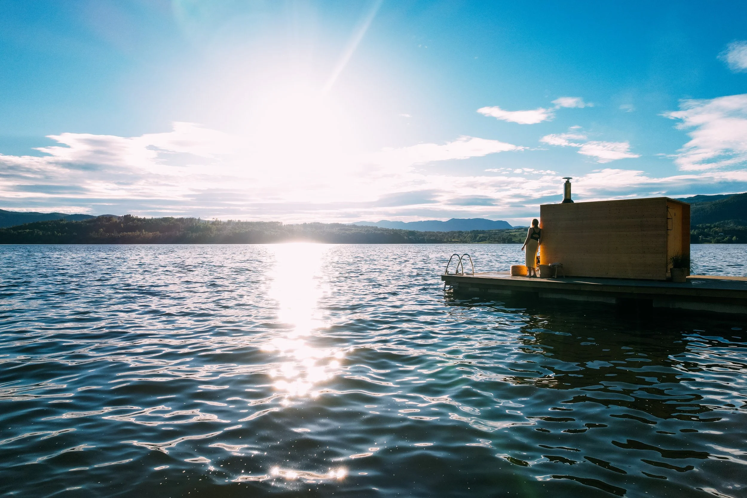 Heit Telemark. En person står på en flytebrygge med utsikt over en innsjø, omgitt av fjell og skyer, med sollys som reflekterer på vannet.