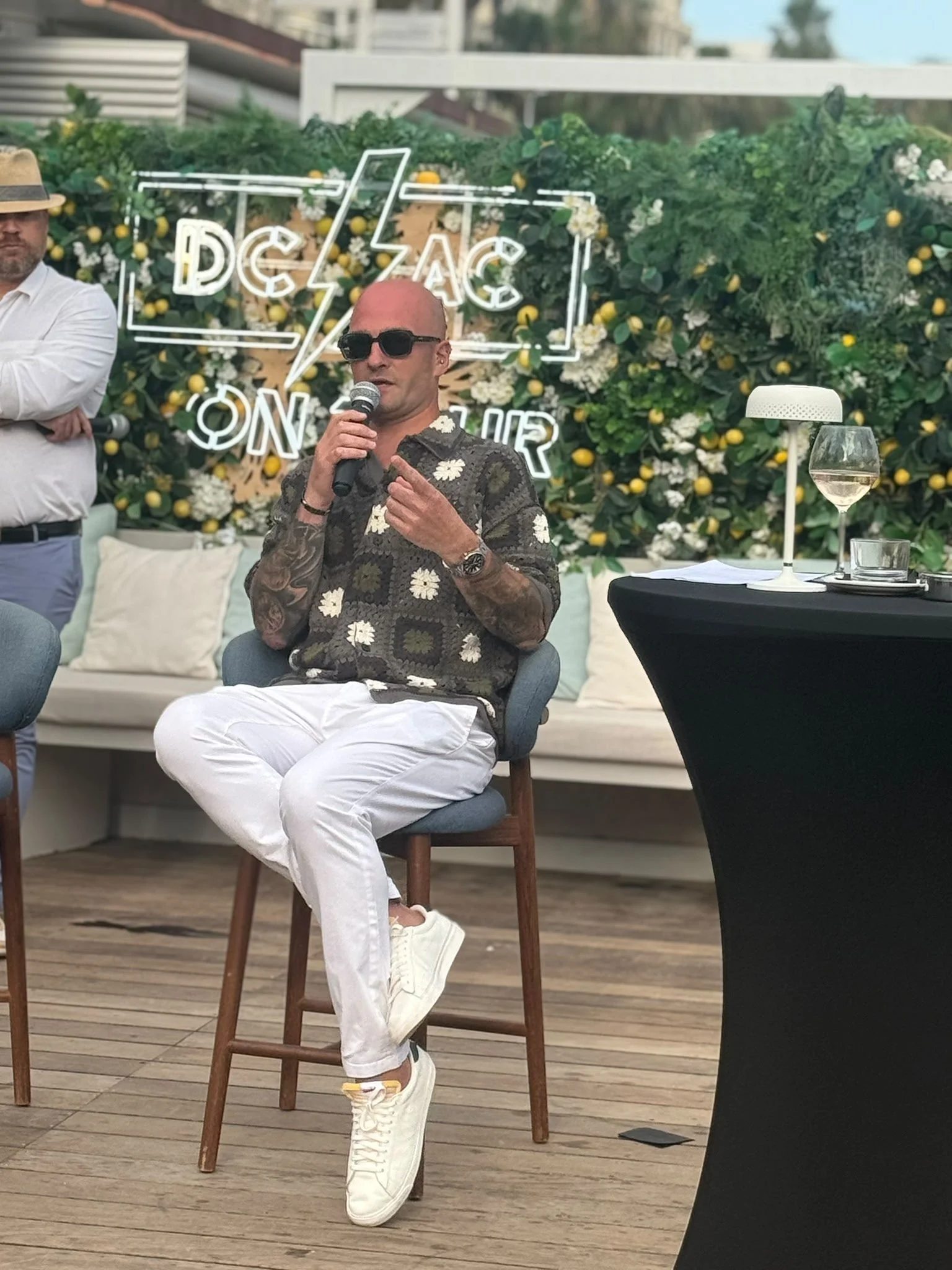 A man in sunglasses wearing a patterned shirt and white pants sitting on a chair, speaking into a microphone at an outdoor event with a flower backdrop and neon sign.