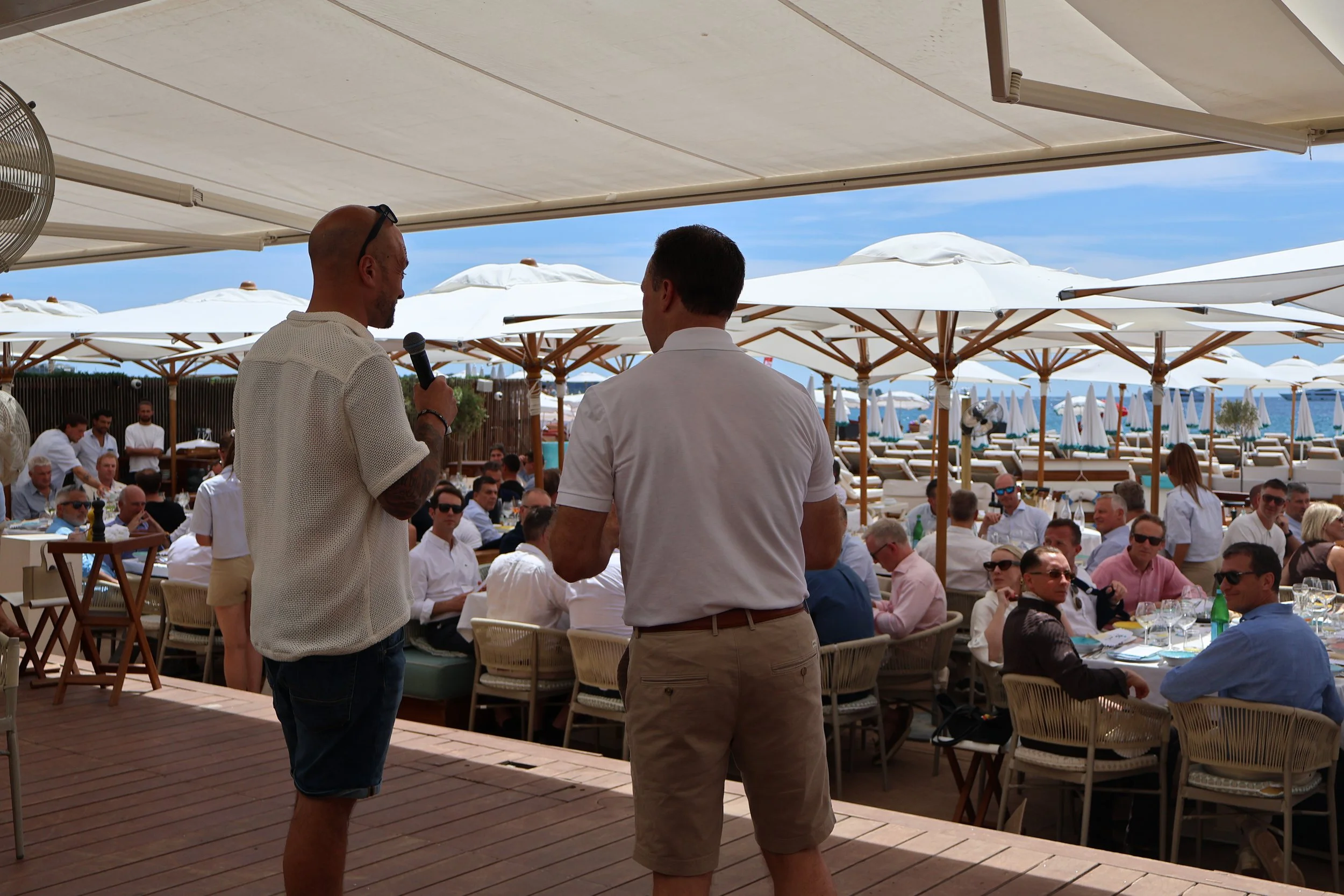 Two men standing and talking at an outdoor restaurant or bar with many people seated and umbrellas in the background.