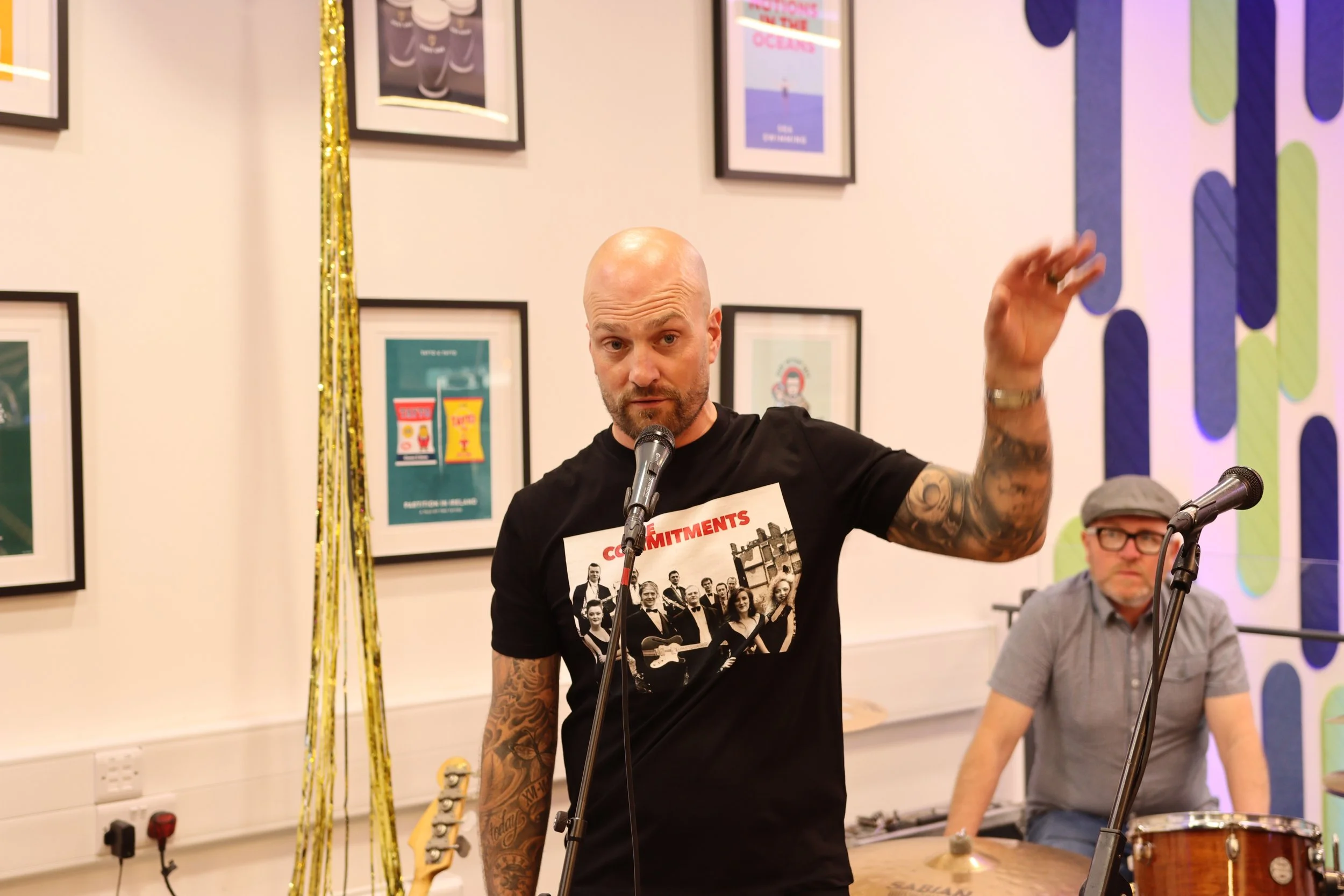 A man with tattoos on his arms speaking into a microphone at an indoor event, with framed posters on the wall behind him. He is wearing a black T-shirt featuring a group photo and text. Another man is seated nearby, wearing glasses and a gray shirt, with a musical drum set visible in the background.