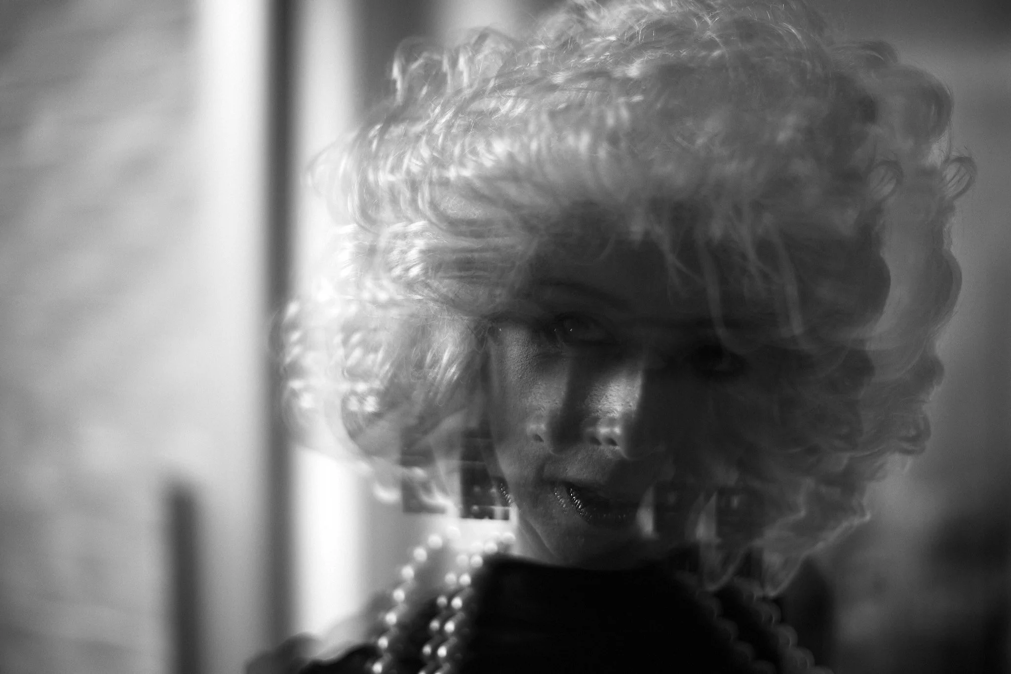 Close-up, black-and-white photo of a person's face and curly hair, partially obscured by a glass surface and other reflections.
