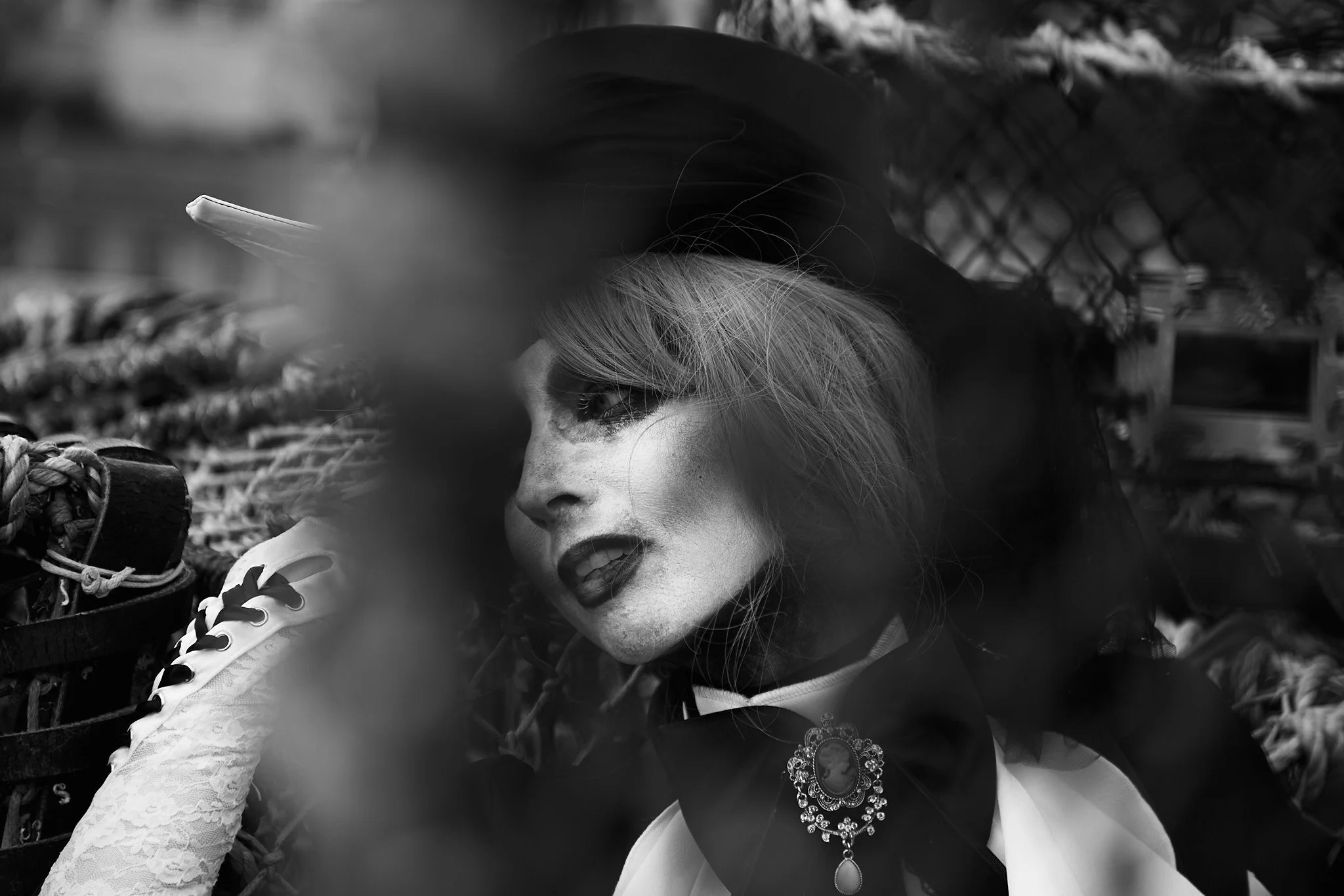 Black and white photo of a woman with blonde hair, lipstick, and makeup, wearing a decorative necklace and lace clothing, lying down with her head resting on her arm, near a chain-link fence with blurred surroundings.