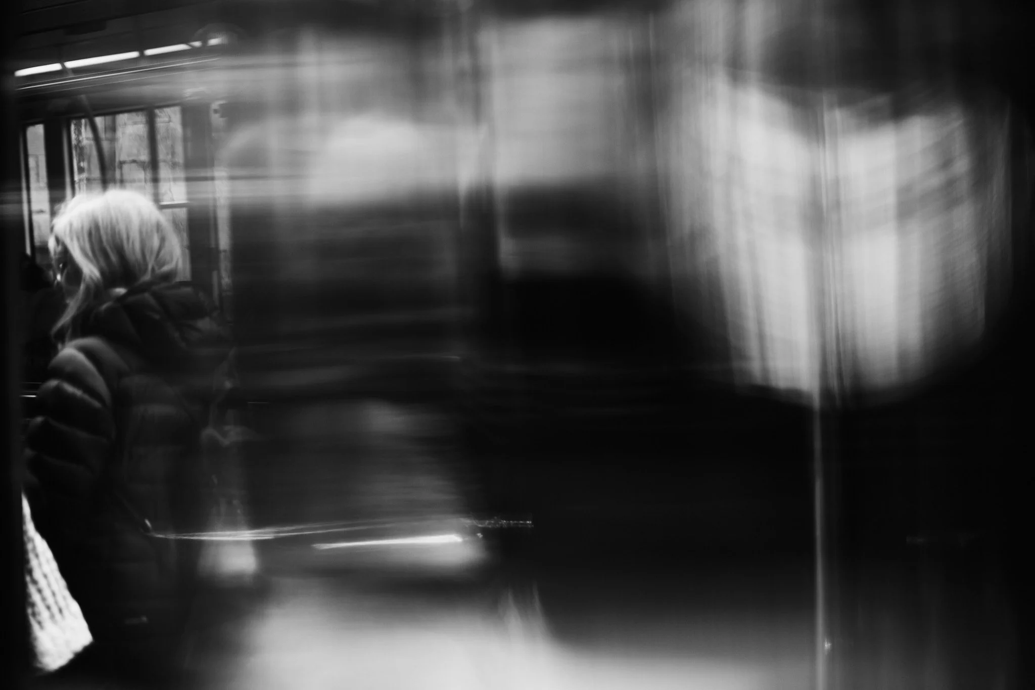 Black and white photo of a person with long hair, wearing a puffer jacket, sitting inside a glass elevator with blurred surroundings.