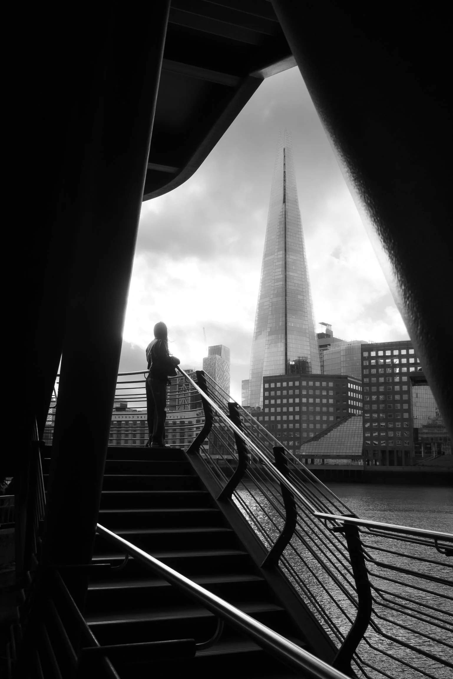 View towards The Shard in London framed by the angular structure of a modern bridge, with a lone figure on the staircase below.