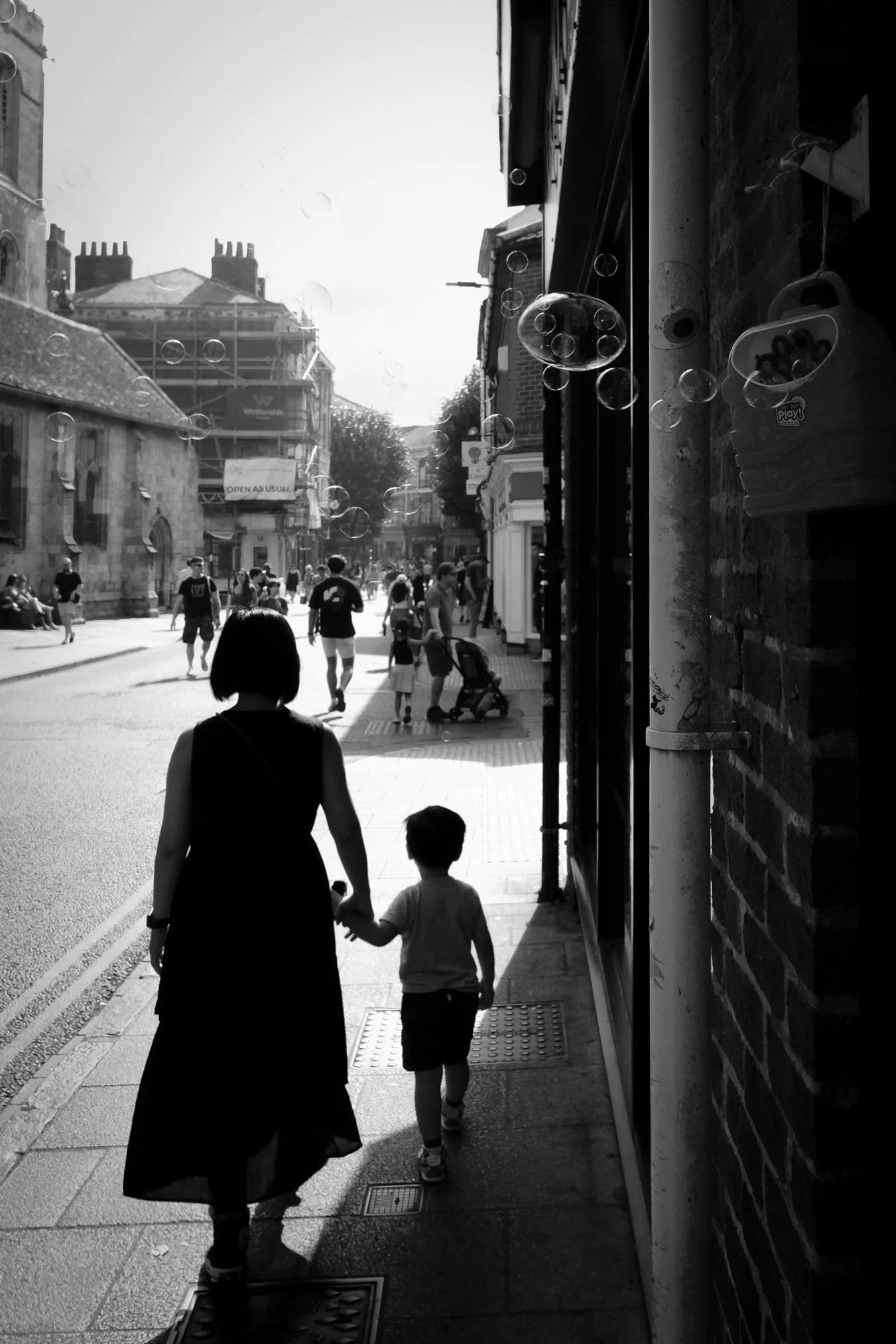 A woman and young child holding hands while walking along a city street, with bubbles floating through the scene.