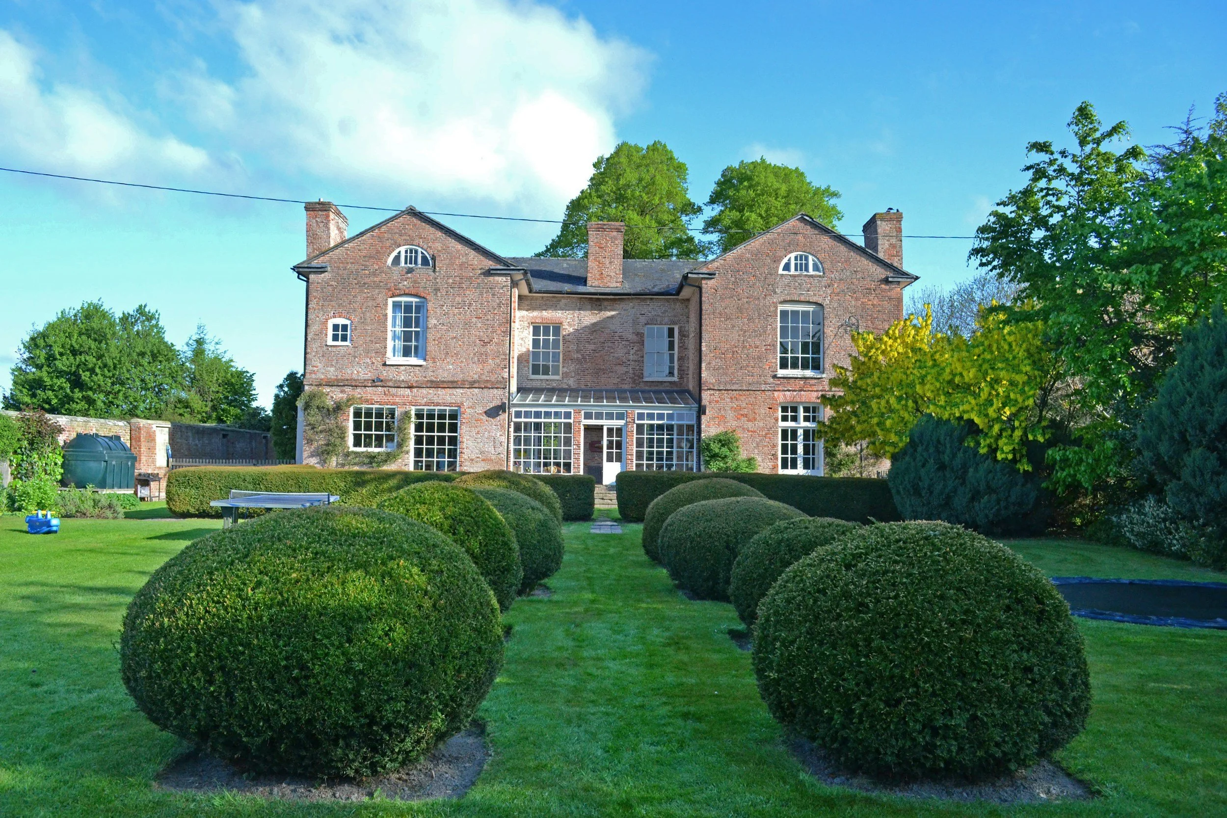 A large brick house with multiple windows, surrounded by a manicured garden with trimmed bushes and lush green trees under a partly cloudy sky.