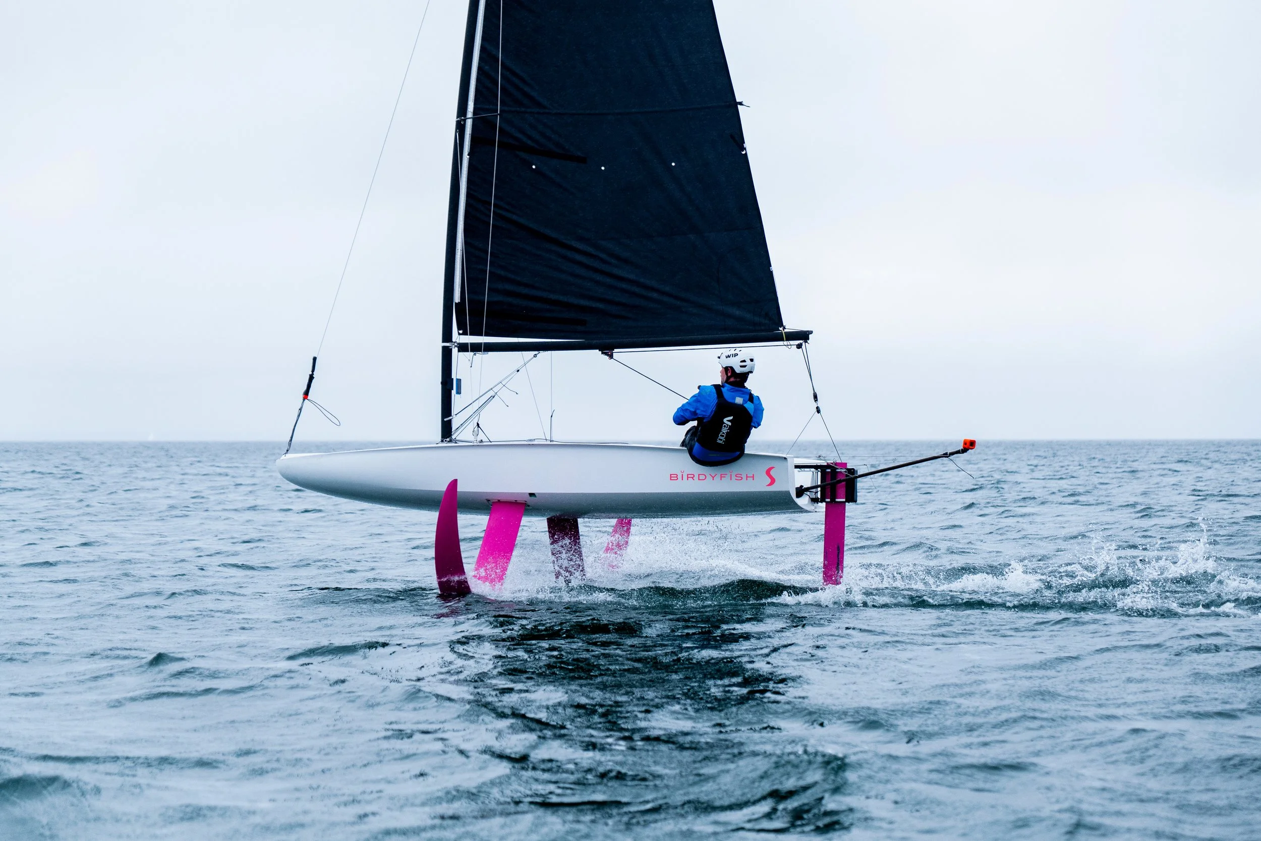 A person sailing a small, black-sailed catamaran with pink hydrofoils on the water, wearing a helmet and life jacket, under an overcast sky.
