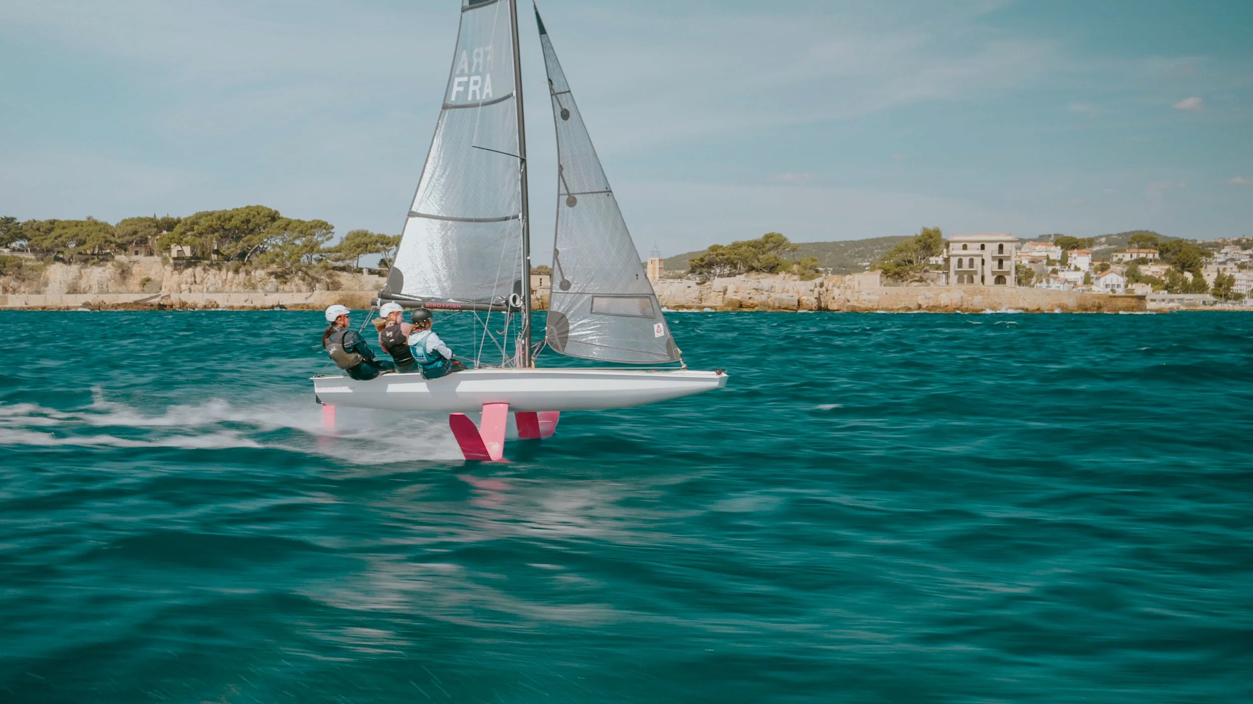 A sailboat with a hydrofoil glides across the water with a coastline featuring trees and buildings in the background.