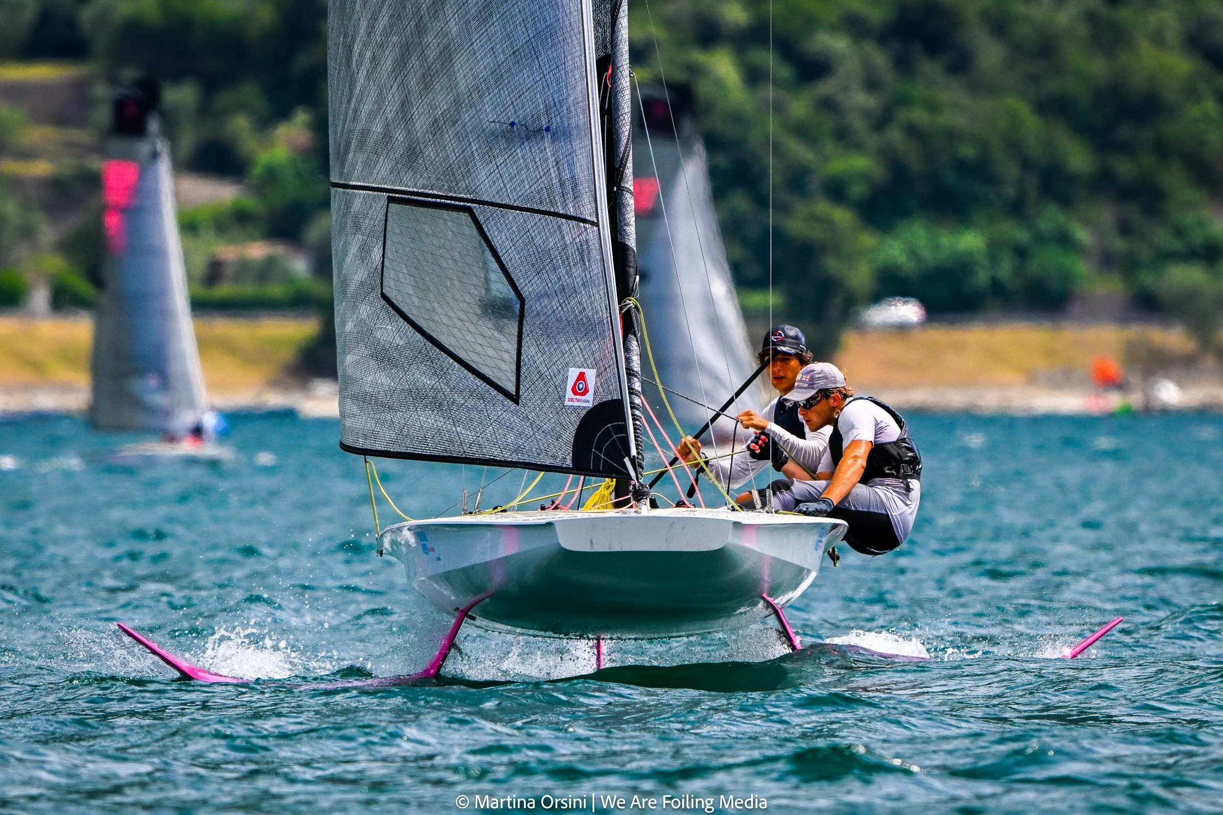 Two sailors on a small racing sailboat with a large, translucent sail navigating choppy water. In the background, there are buoys and lush green trees along the shoreline.