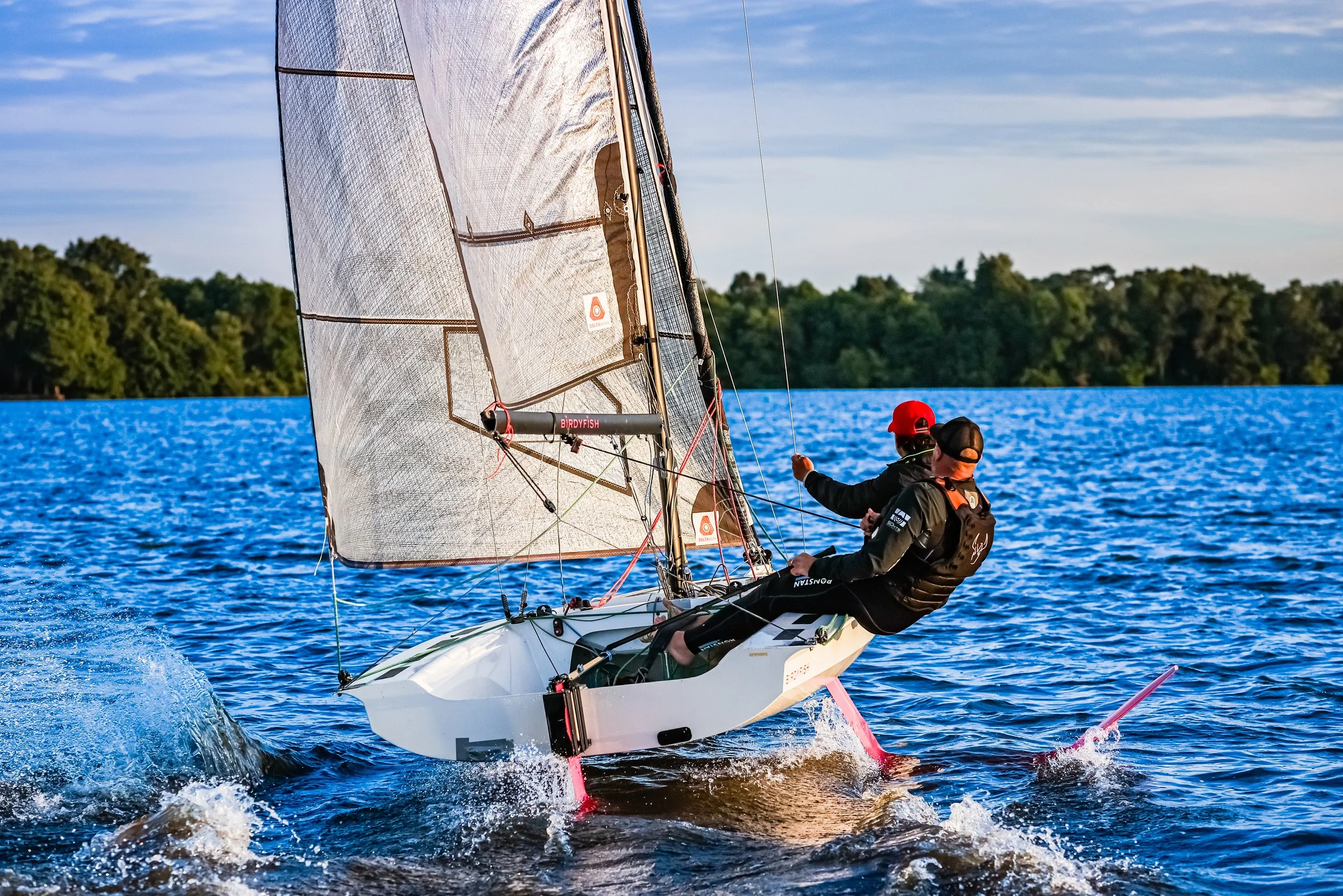 Two sailors in black jackets and red and black caps sailing a small racing boat with a large sail on a blue body of water, with a green tree-lined shoreline in the background and a clear sky above.