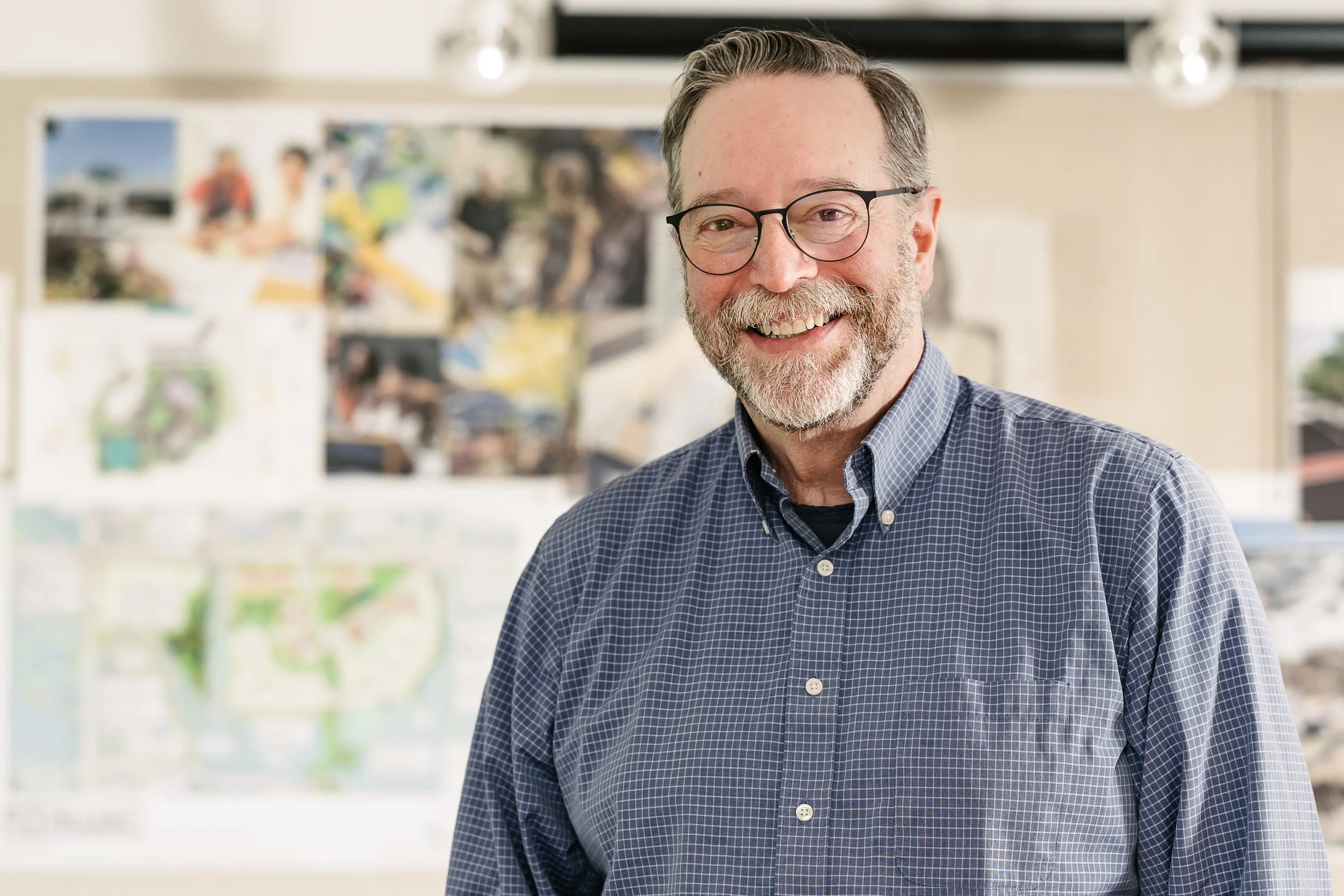 Lee Brown wearing a blue checkered shirt in a bright interior space with blurred artwork or posters in the background.