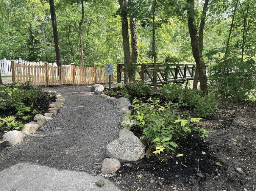 A gravel pathway in a wooded park area, bordered by rocks, leading to a small wooden bridge and a wooden fence.