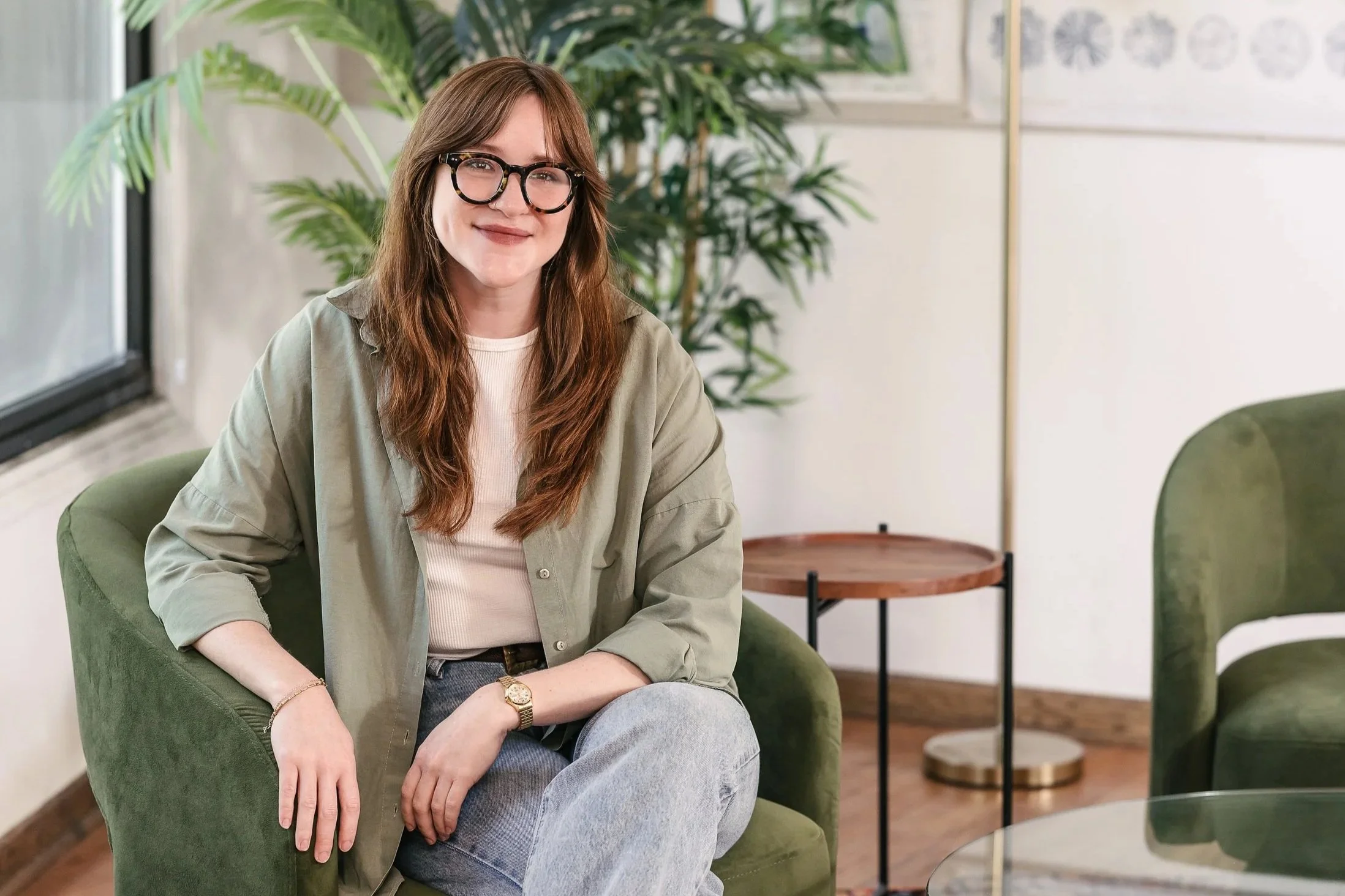 Maggie Eickhoff sitting on a green chair in a room with large windows, plants, and minimalist decor.