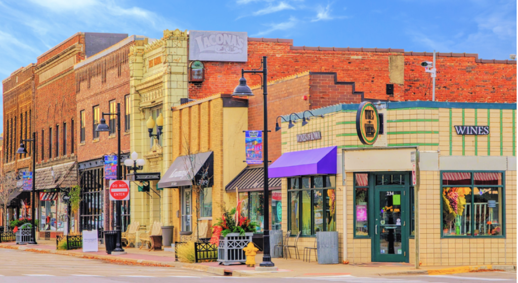 A street view of Historic Valley Junction in West Des Moines, Iowa showing small businesses with colorful buildings, storefronts, hanging street lights, and flower displays in a downtown area on a bright day.
