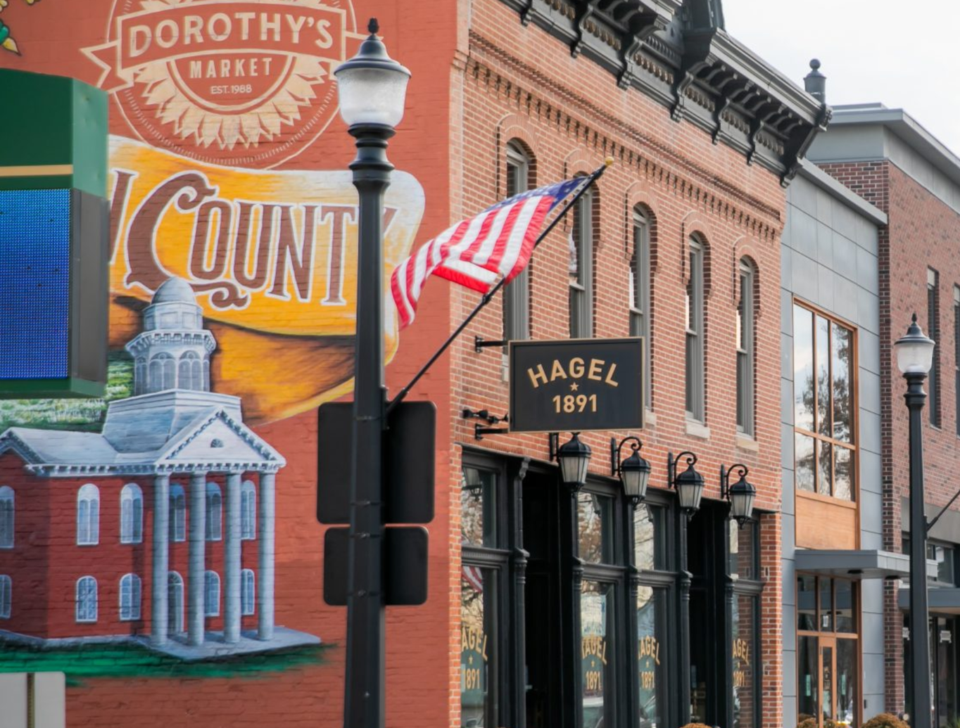 Street view of a brick building with large windows, decorative lamp posts, and a mural painted on the wall.