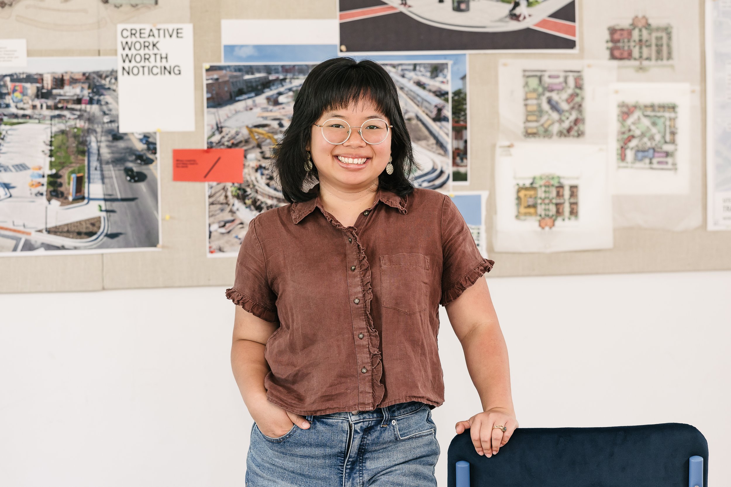 Maly Sears stands in front of a bulletin board with various architectural and aerial photos, smiling at the camera.