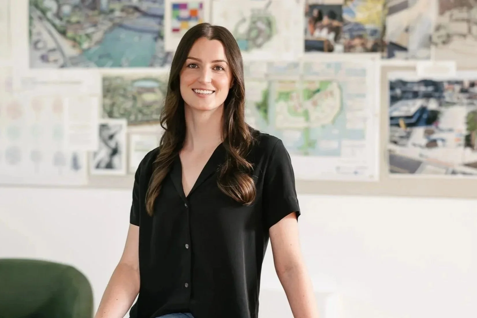 Emma Swanson wearing a black shirt, smiling at the camera in an office or classroom with maps or plans on the wall behind her.