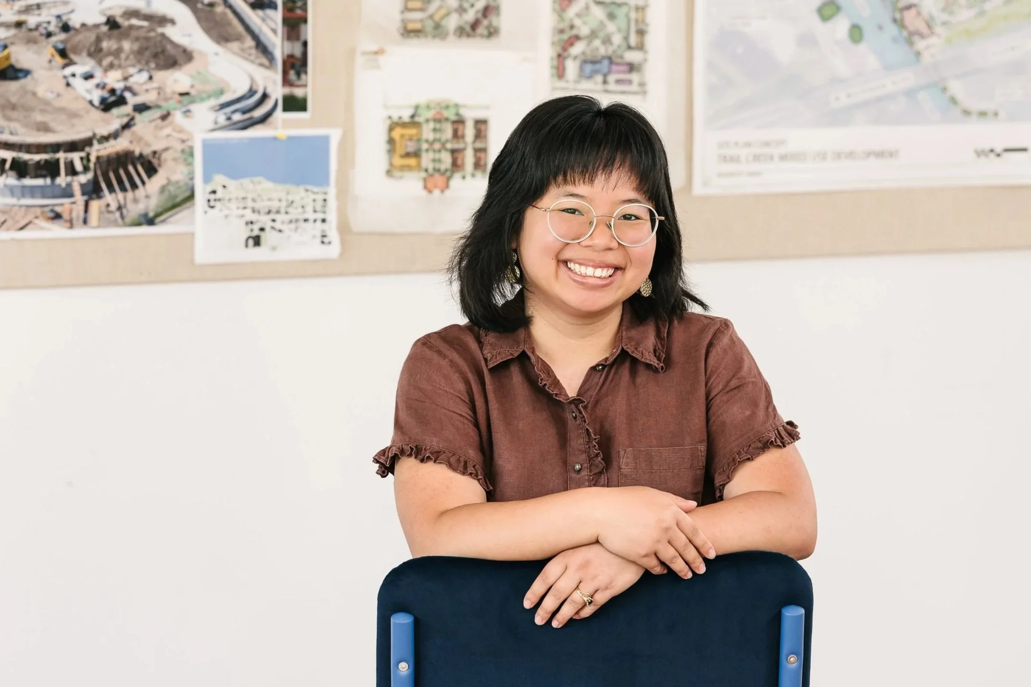 Maly Sears smiling while sitting with her arms crossed, in front of a beige wall with architectural plans and images pinned to it.