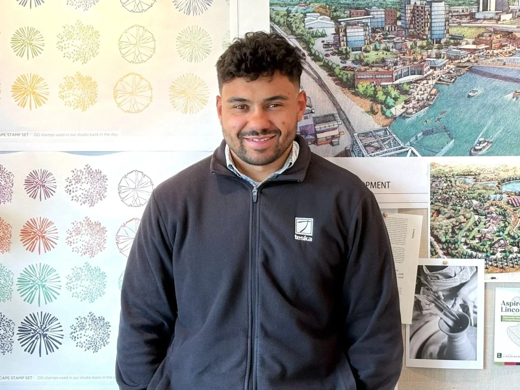 Aaron Lewis smiling and standing in front of a colorful mural and posters relating to cityscape and ocean activities, wearing a black jacket with a logo on it.