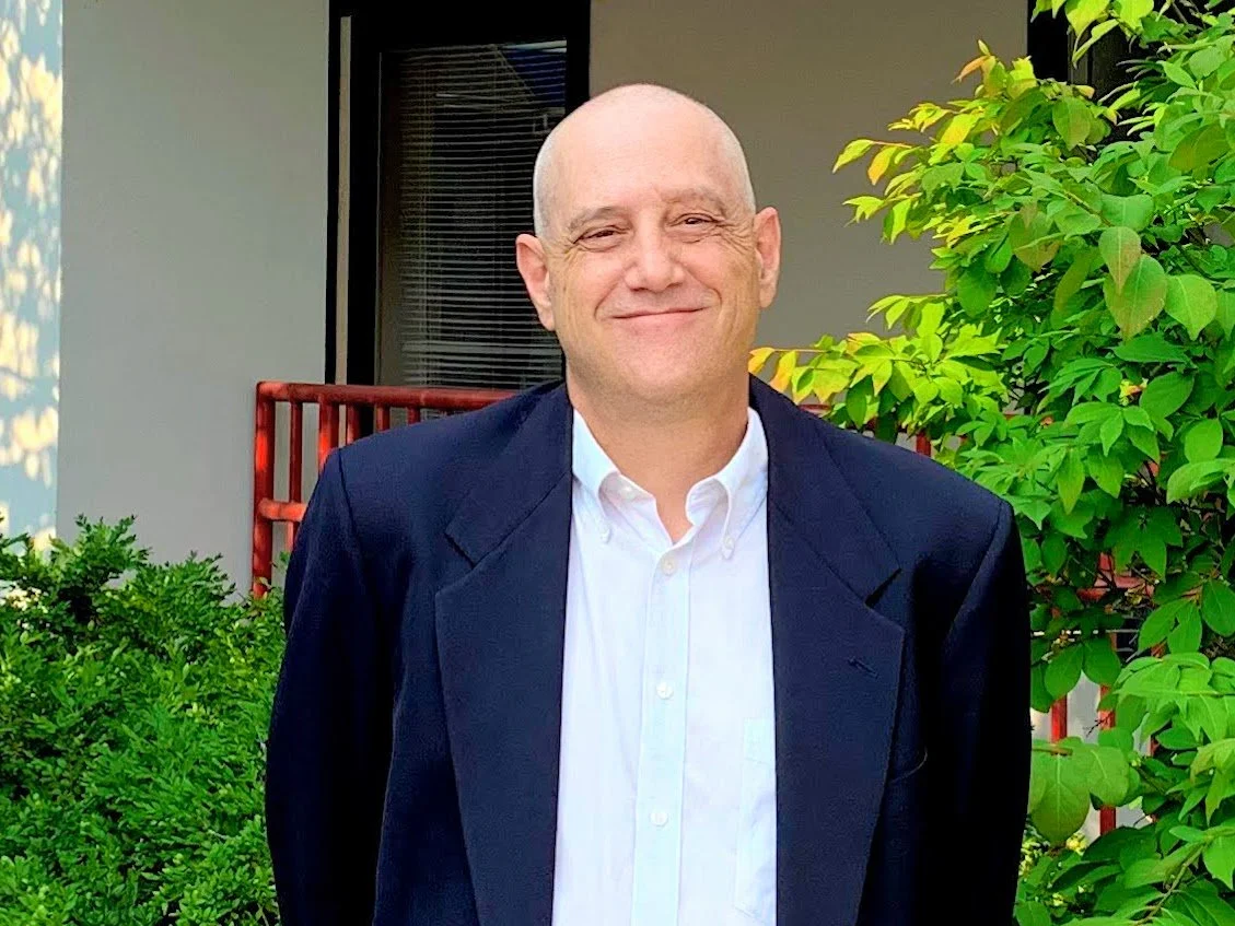 Pete Iosue wearing a dark suit jacket and white dress shirt standing outdoors near green foliage and a building with a window.