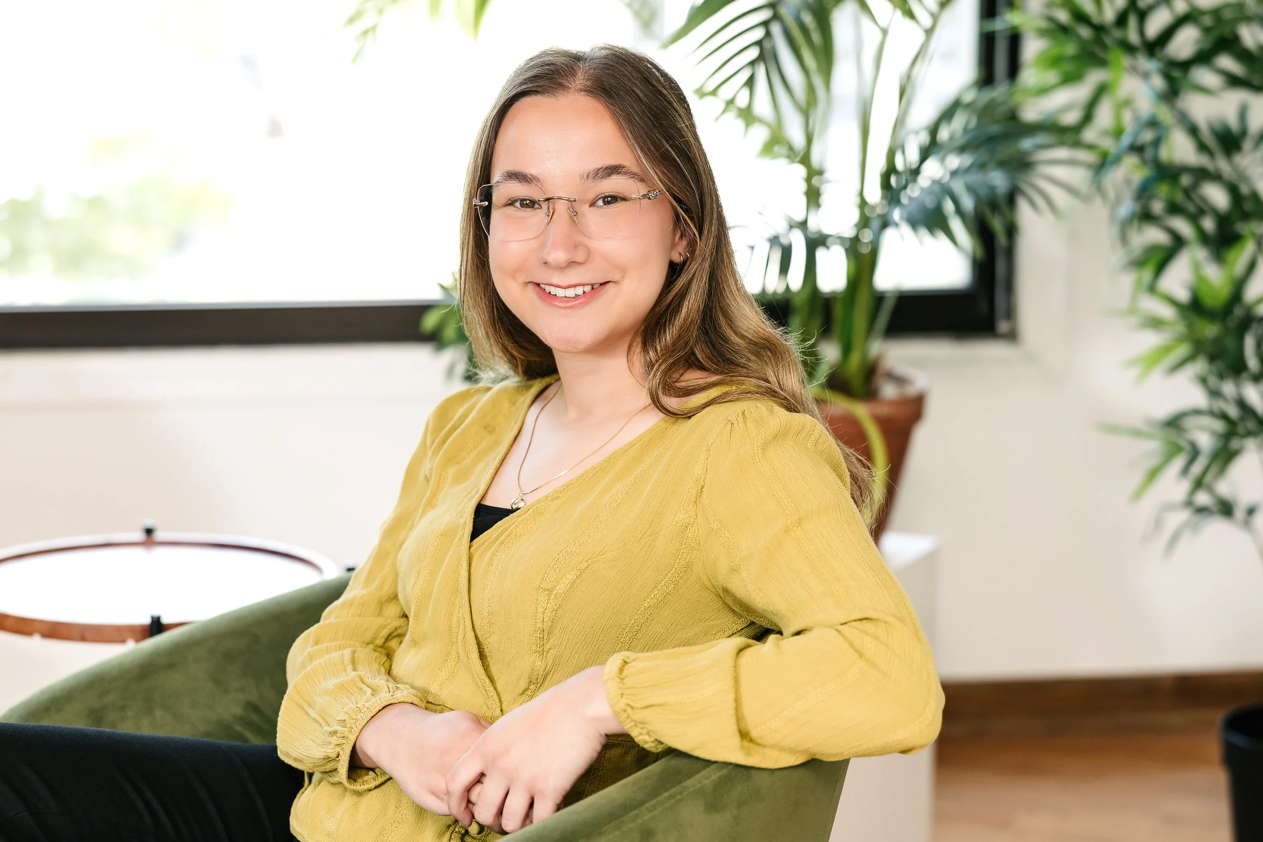 Natalie Crown sitting on a green chair inside a room with large windows and tropical plants in the background.