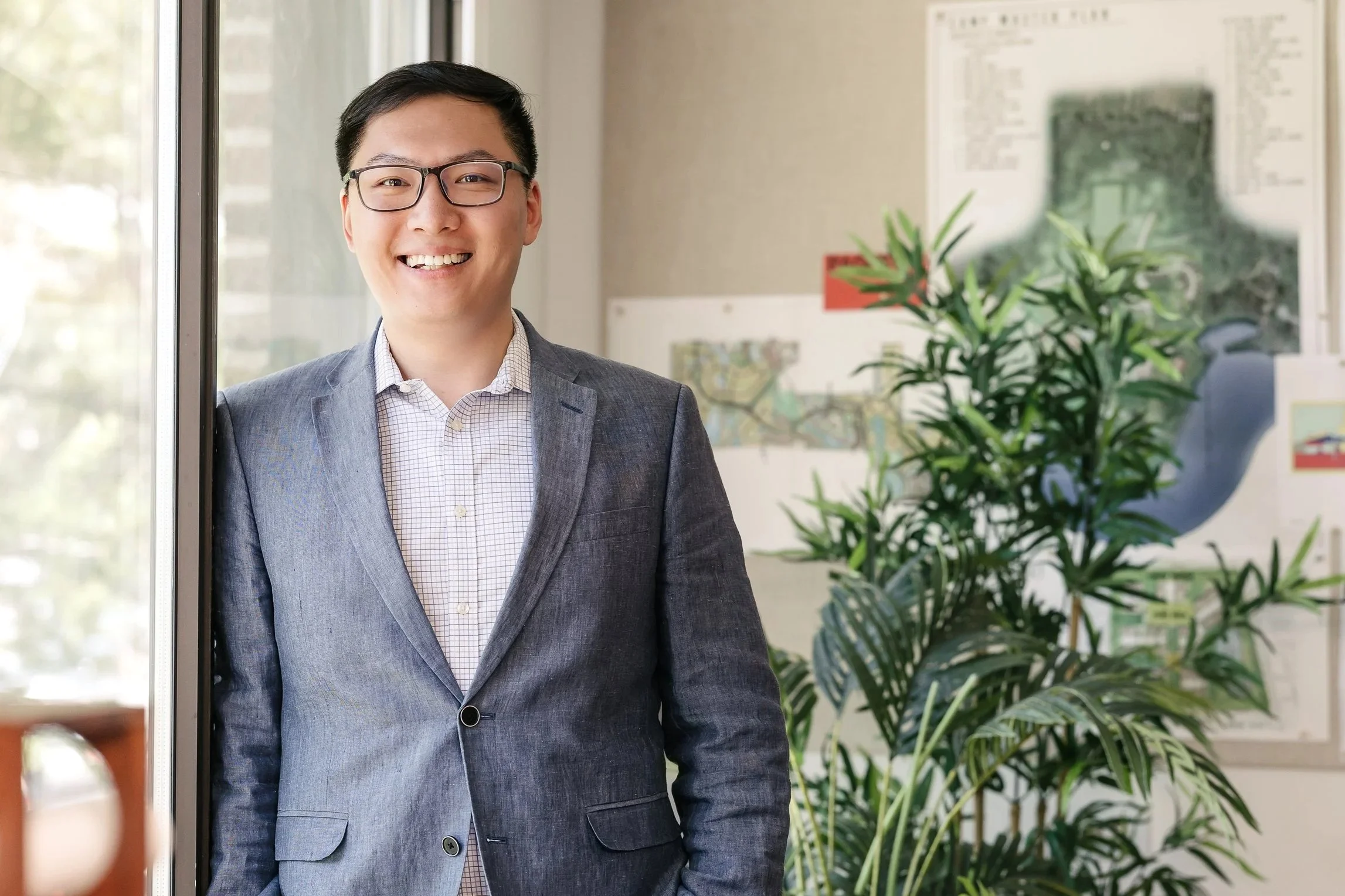 Yuchen Ding standing in an indoor office space near a window, with plants and maps visible on the wall behind him.