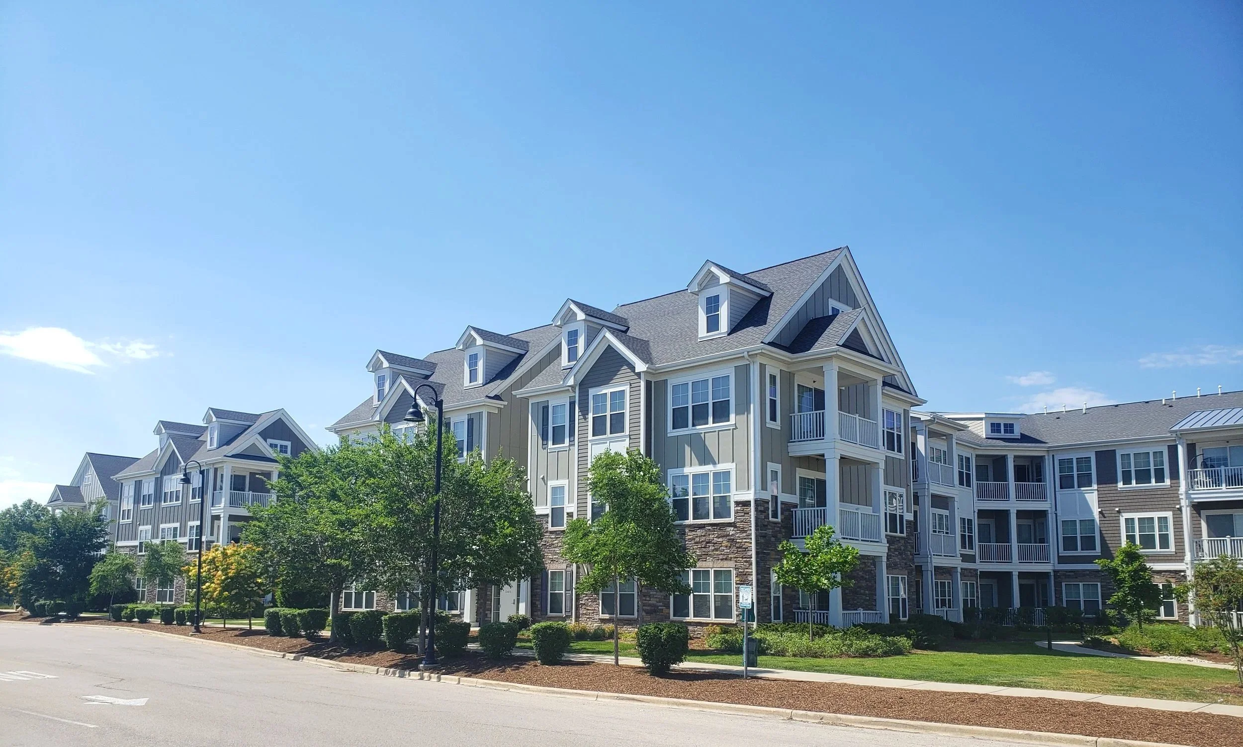 A row of modern multi-story apartment buildings with gray siding and white trim, surrounded by trees and a clear blue sky.
