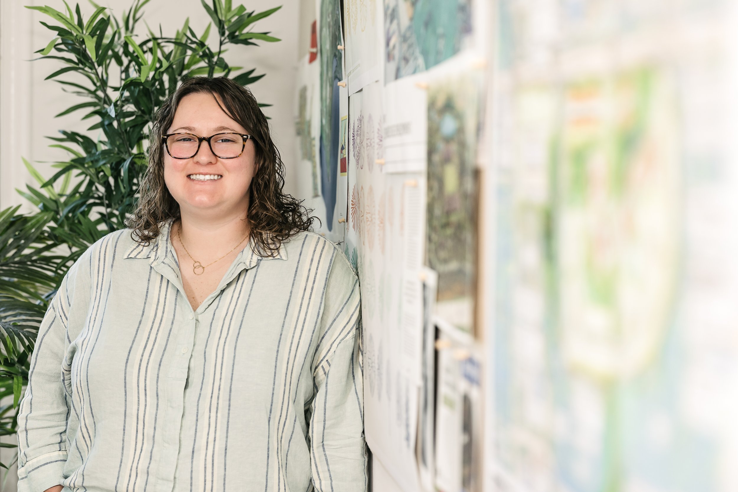 Aliza Courey woman with glasses and curly hair stands in front of an arts and crafts display wall, smiling and wearing a striped shirt.
