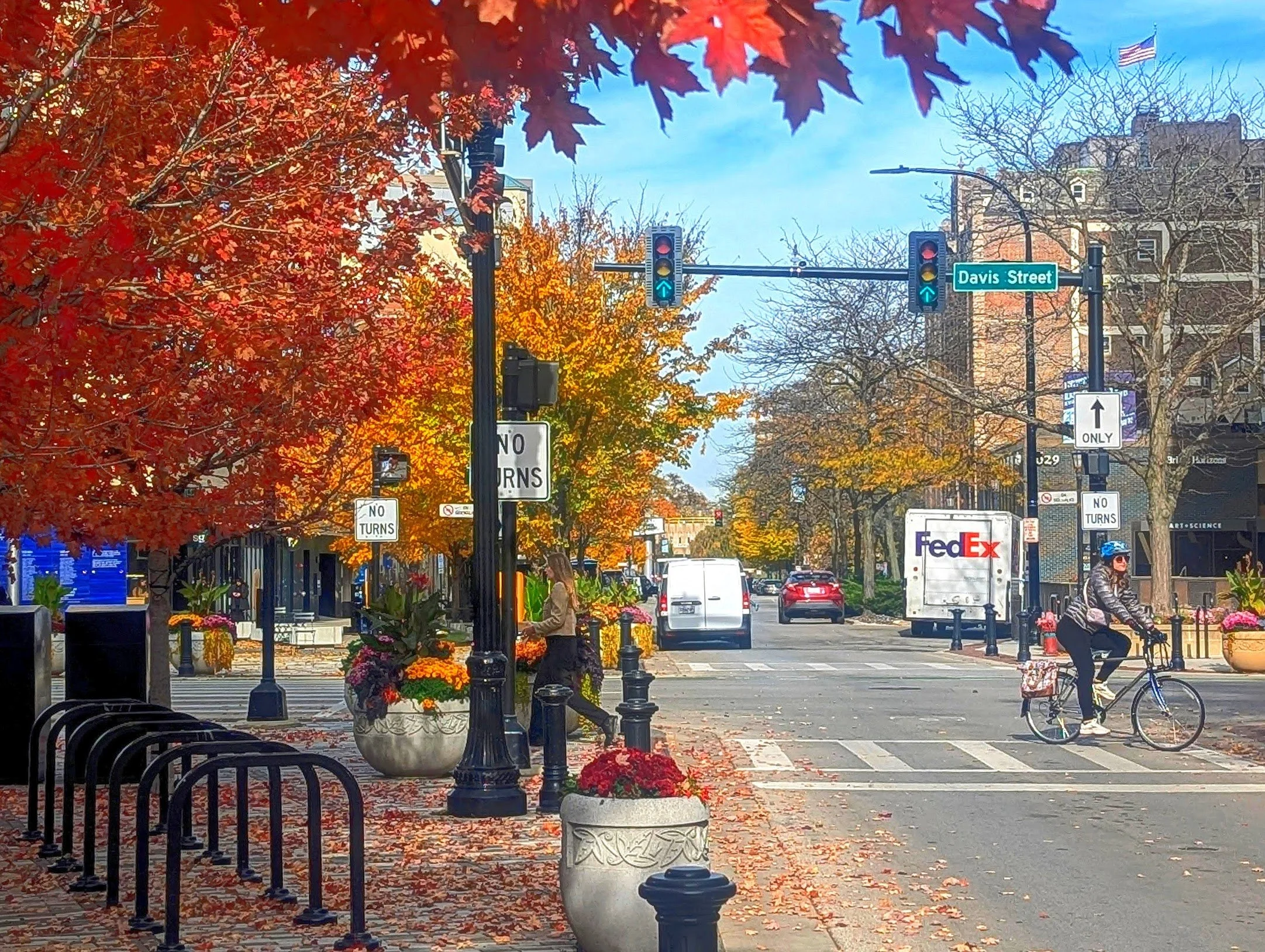 Evanston street scene in autumn, featuring colorful fall foliage on trees, a crosswalk, pedestrians, and vehicles. Decorative planter pots with flowers are also visible.