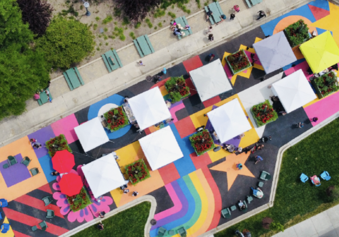 Aerial view of a colorful outdoor marketplace with white tents, flower boxes, and rainbow-themed painted walkways.