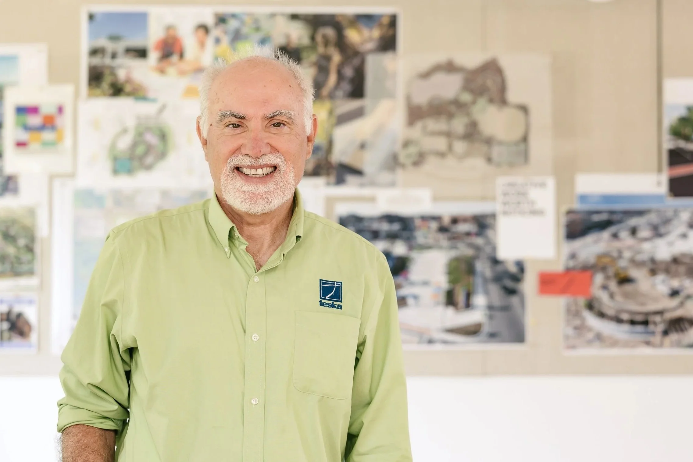 Mike Hoffman wearing a light green shirt with a logo that says 'Teska,' standing in a room with blurred architectural drawings or blueprints on the wall behind him.