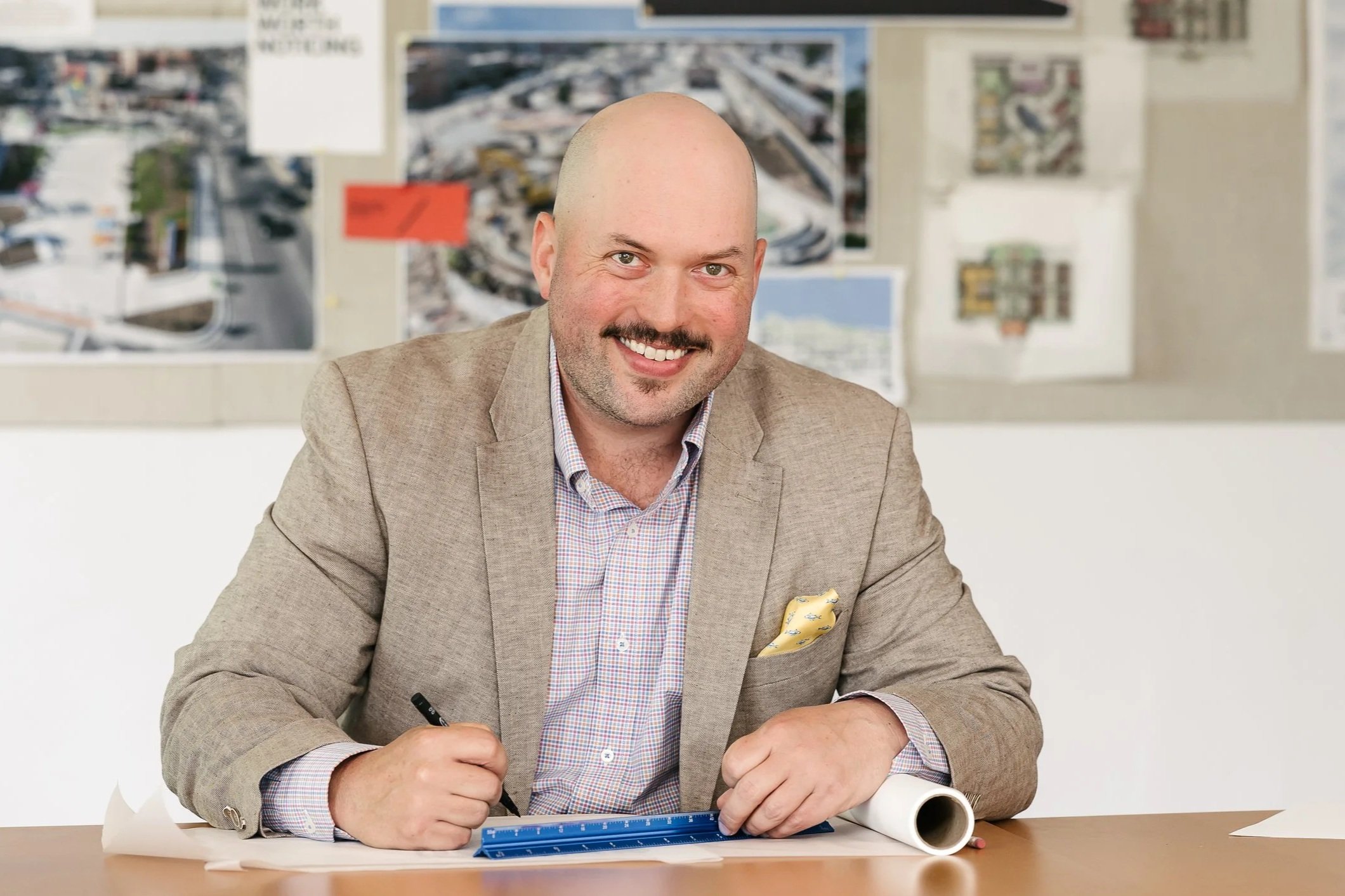 Danny O'Brian sitting at a desk with architectural plans and a ruler, in an office with place maps on the wall behind him.