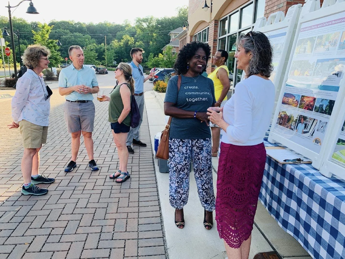 Group of people outdoor at community event, some engaged in conversation near informational display boards on tables with checkered tablecloth.