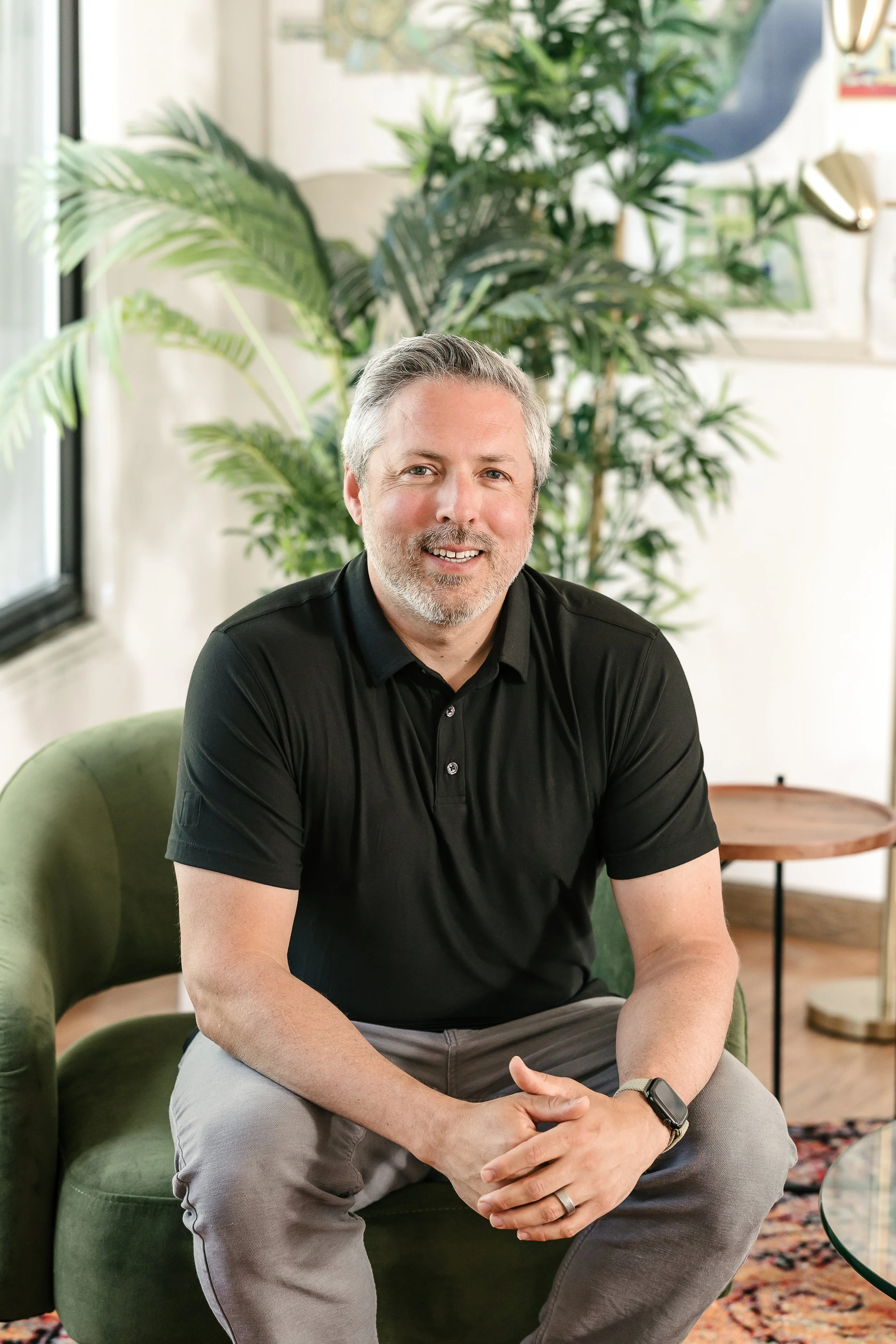 Dominic sits on a green armchair in a bright room. Behind him are large green plants, a small wooden side table, and a patterned rug.