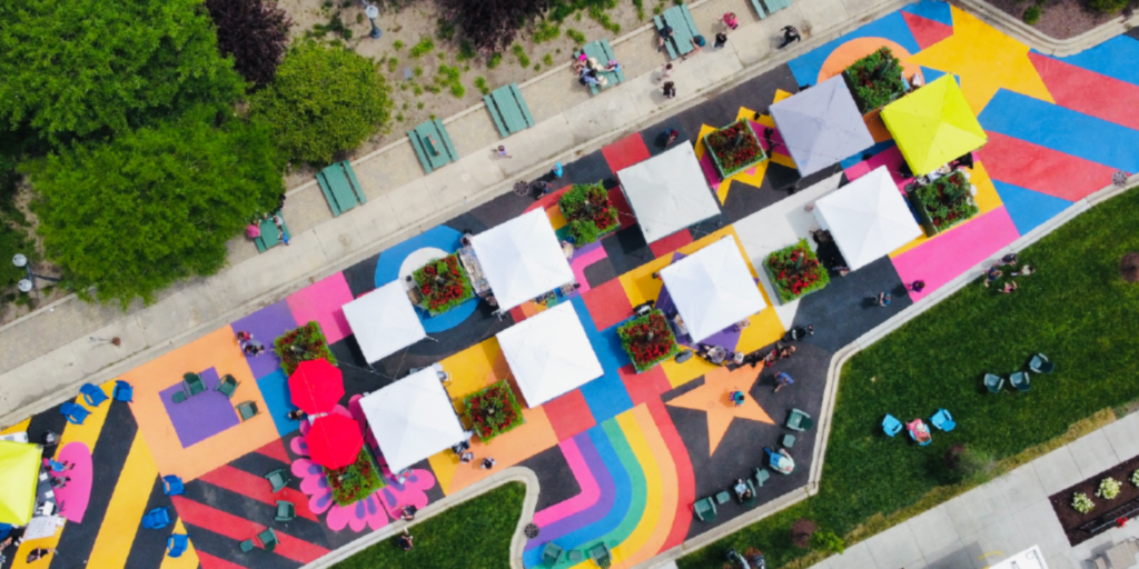 An aerial view of a colorful outdoor market or event with white tents on a vibrant rainbow-themed pavement, surrounded by green grass and trees.