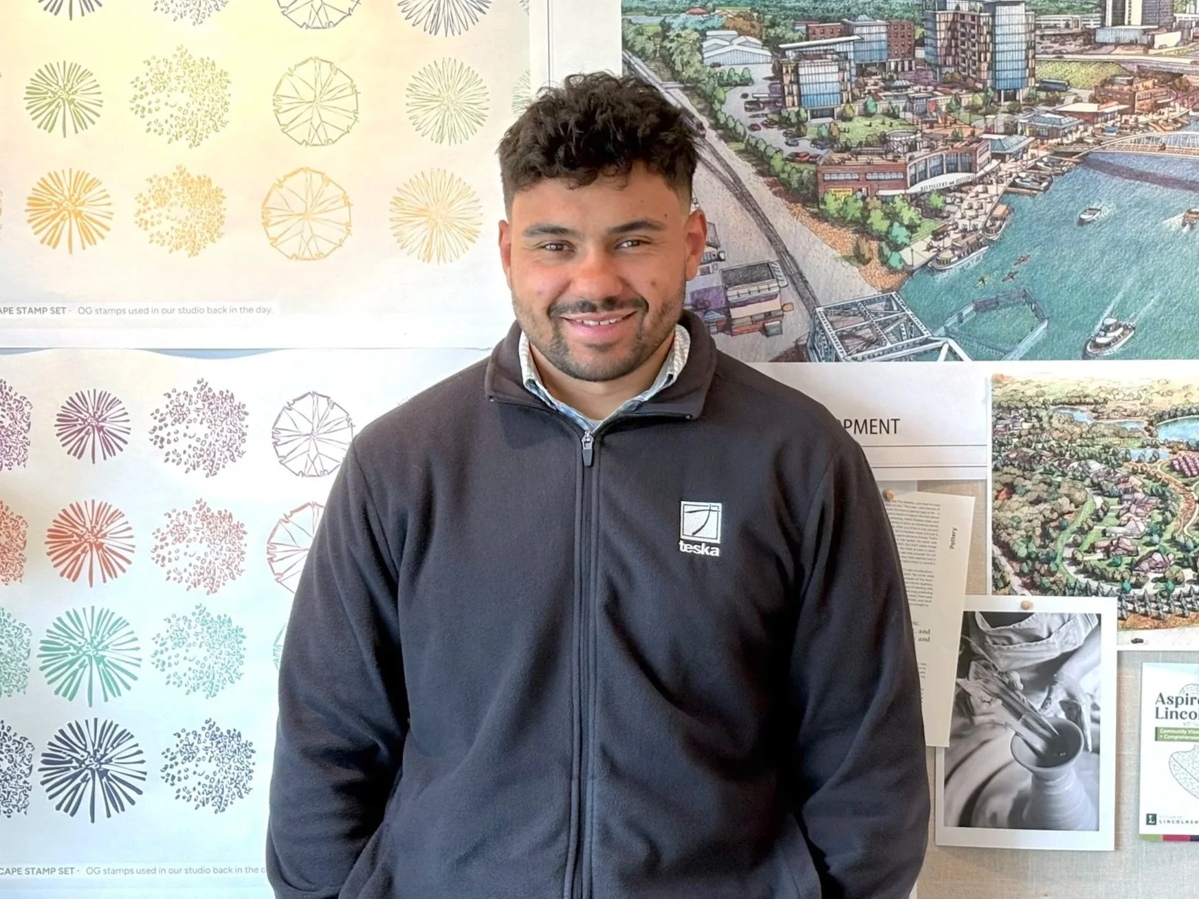 Aaron Lewis smiling, wearing a black jacket with a logo and a collared shirt, standing in front of a colorful urban development illustration and decorative wall art.