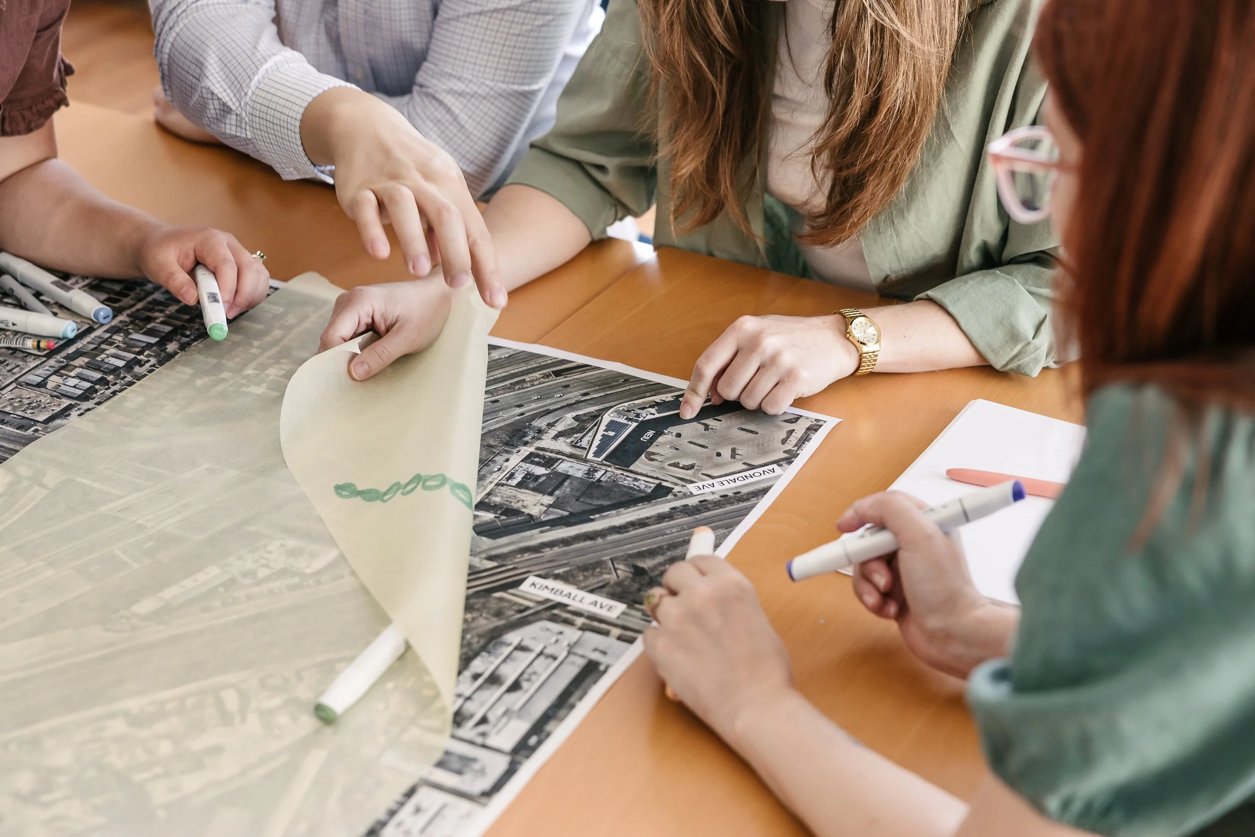 Multiple people gathered around a table examining a black and white aerial map with street names and landmarks, discussing planning or development.