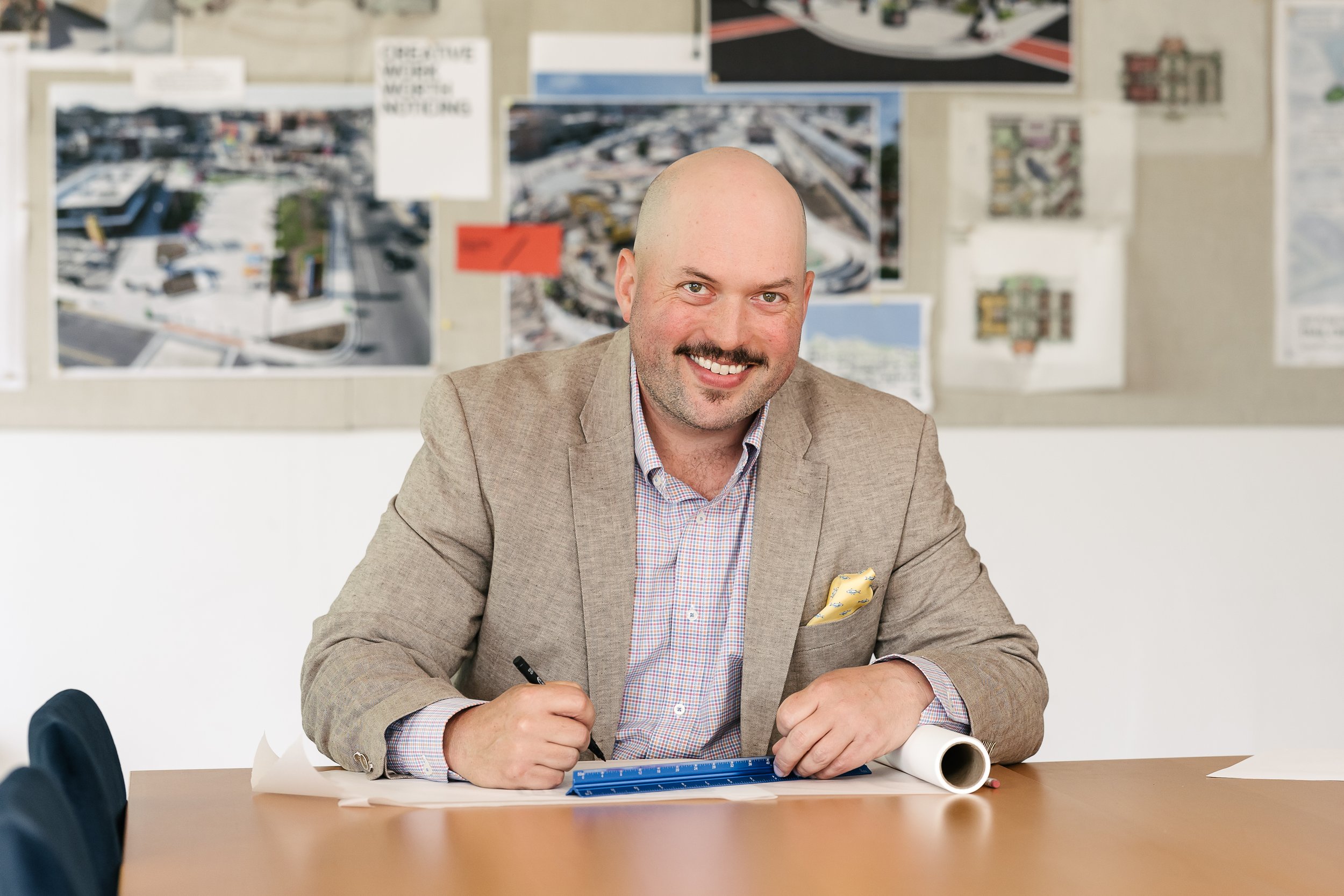 Danny O'Brian wearing a beige blazer and a plaid shirt, is smiling while sitting at a table in a room with maps and plans on the wall behind him.