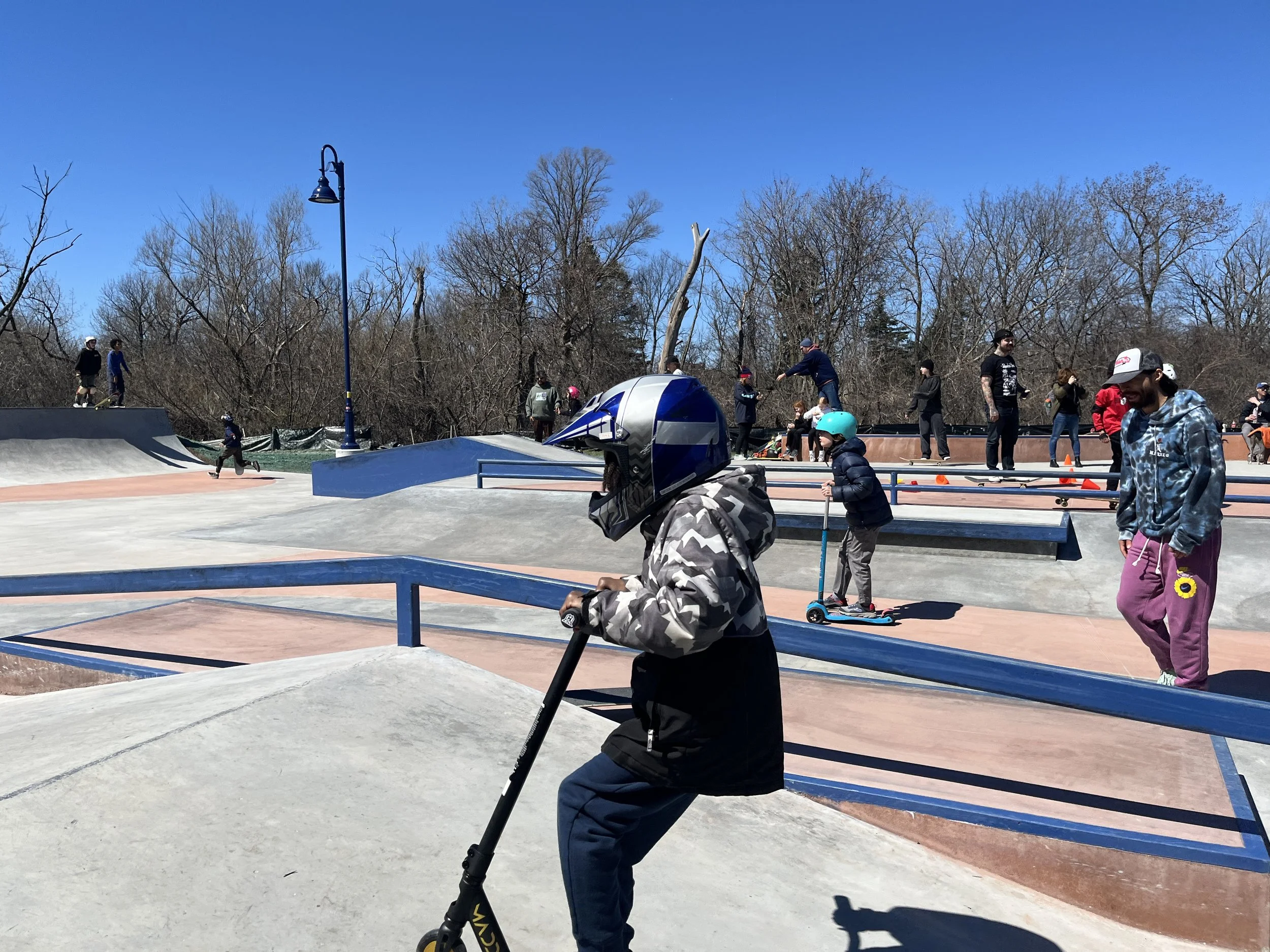 People enjoying skateboarding at Evanston skate park on a sunny day, with leafless trees and a clear blue sky in the background.