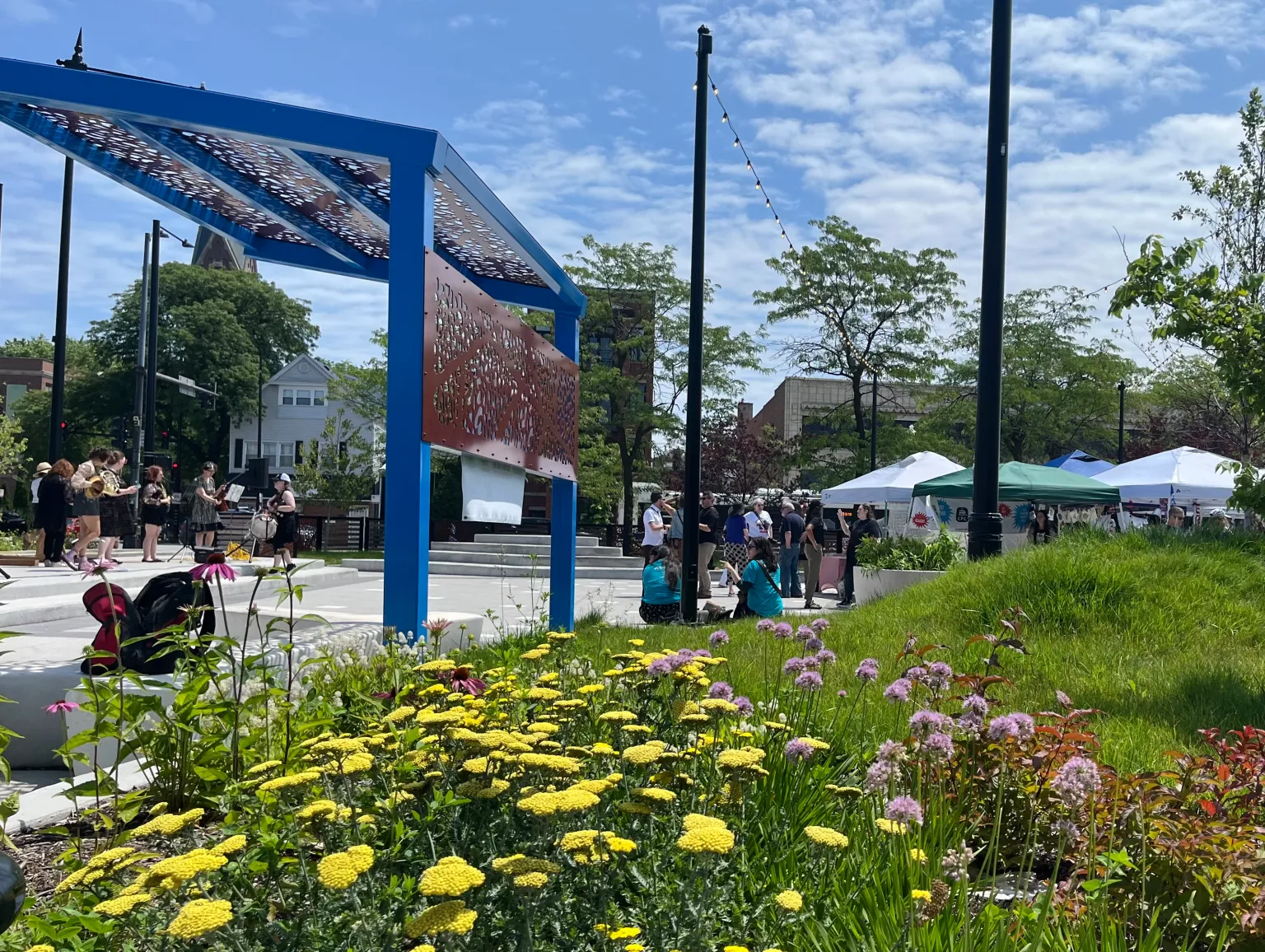People gathering at an outdoor plaza in Lincoln Square with tents, flowers, and a blue informational kiosk on a sunny day.