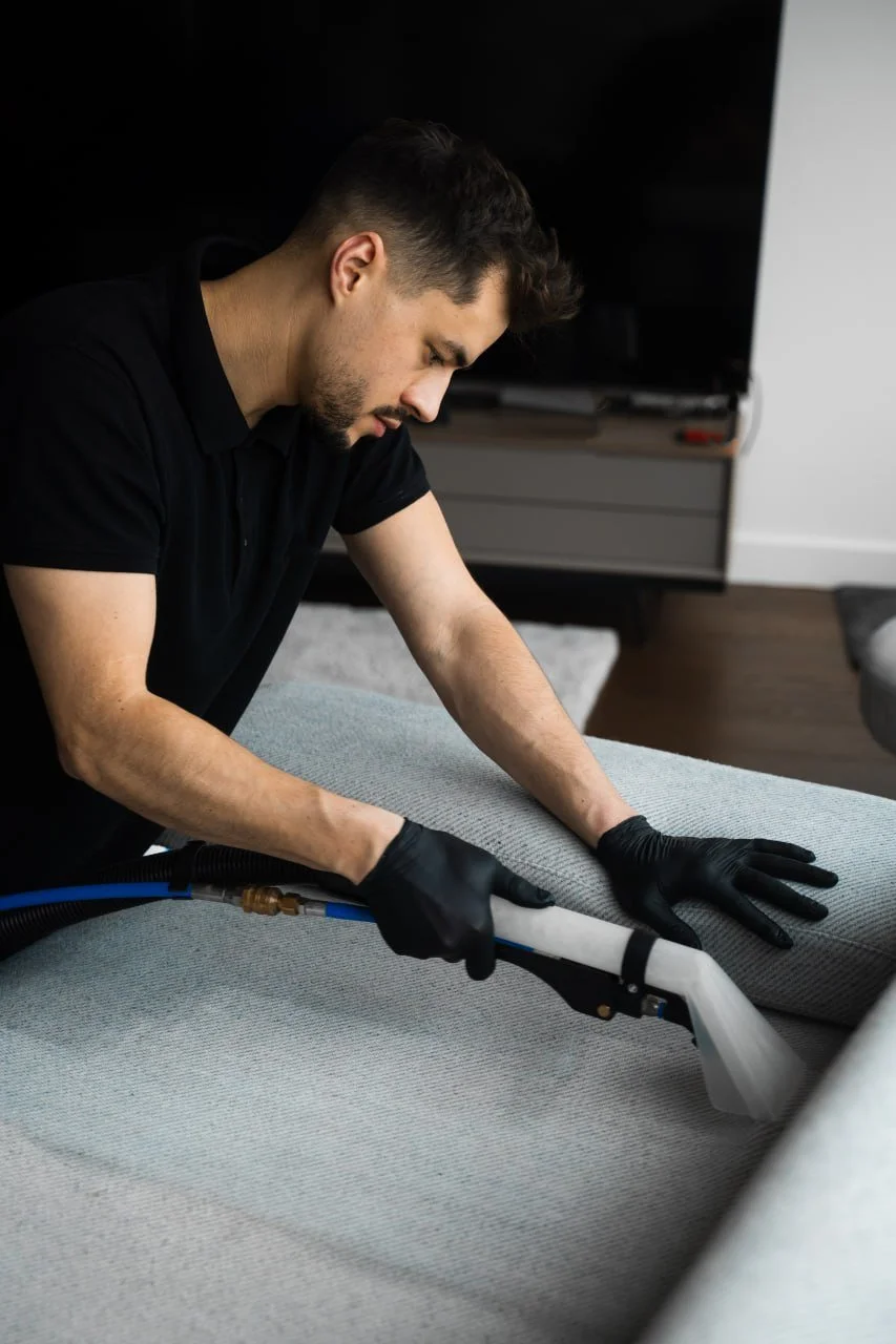 Man cleaning or disinfecting a grey upholstered sofa with a spray gun, wearing black gloves.