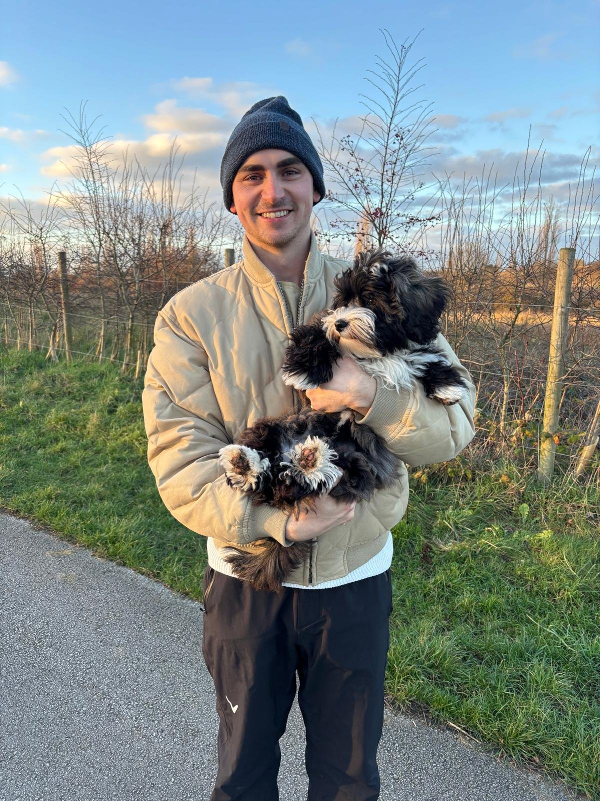 A young man with a beanie hat and beige jacket holding a black and white puppy outdoors near a fence and trees, with a blue sky and clouds in the background.
